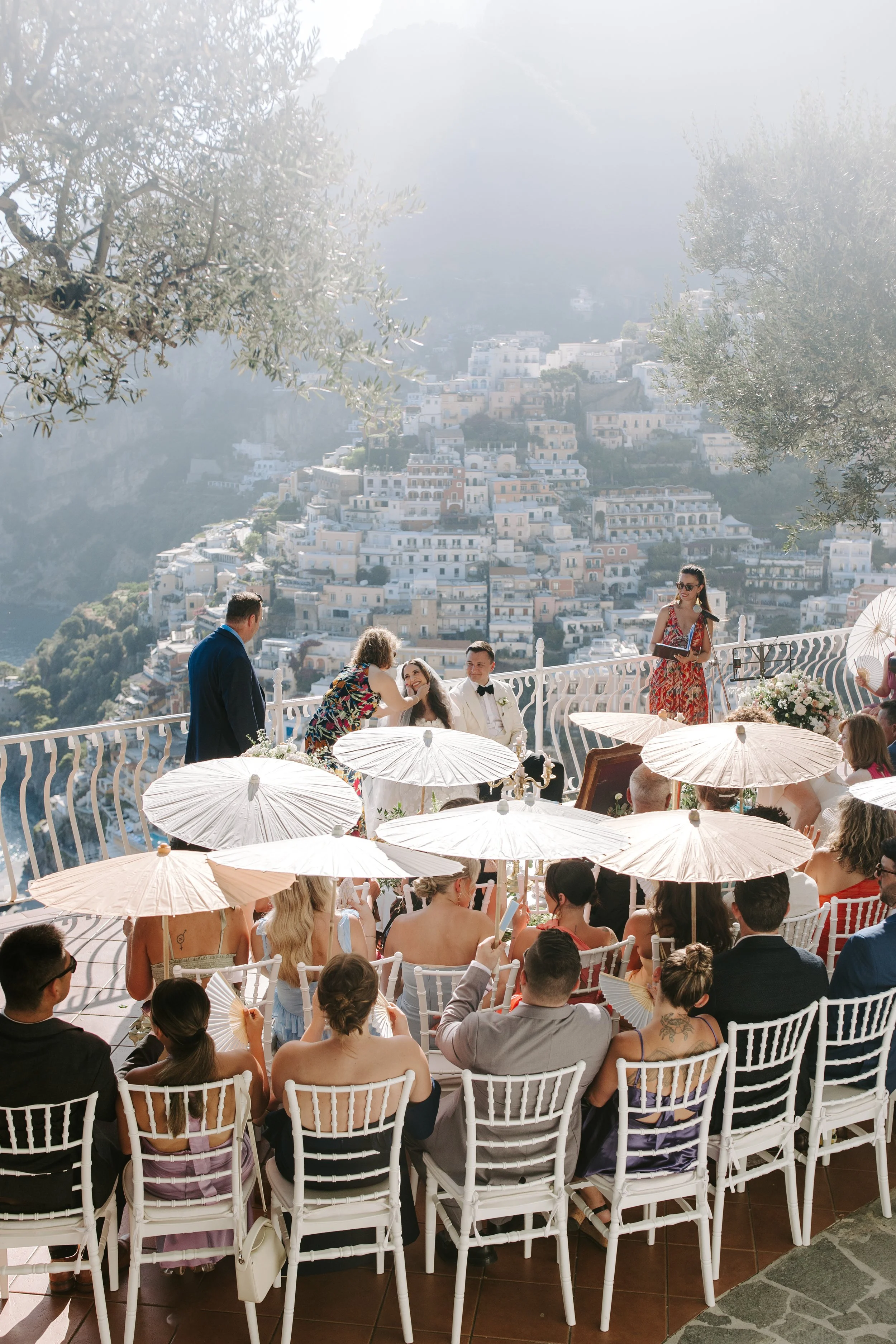 Wedding ceremony on a terrace with a view of a hillside town, guests holding white fans, and a woman in a red dress speaking, with a decorated table nearby.
