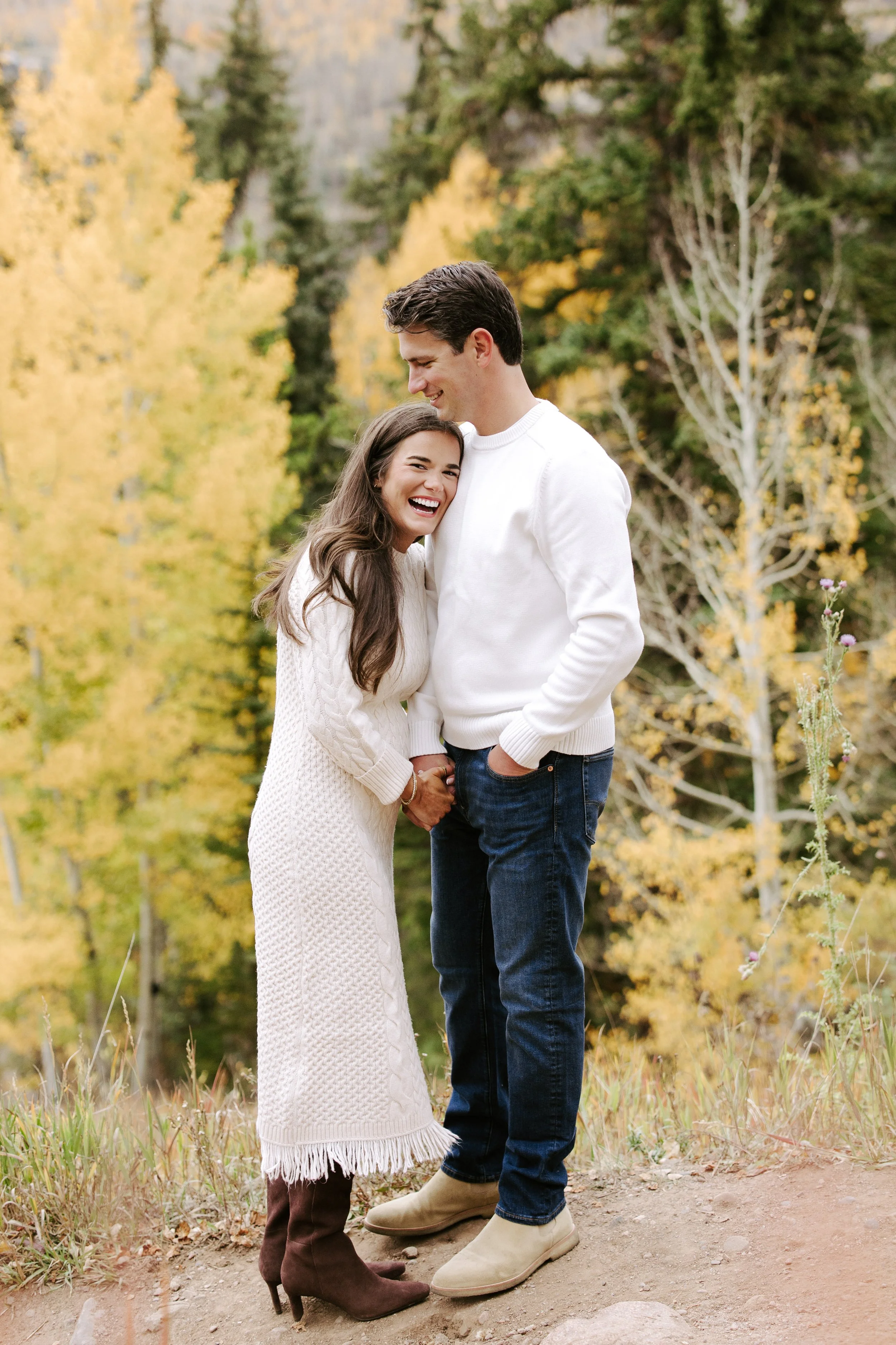 A young couple standing outdoors in autumn, smiling and holding hands. The woman wears a white sweater and brown boots, and the man wears a white sweater and beige boots. There are yellow and green trees in the background.