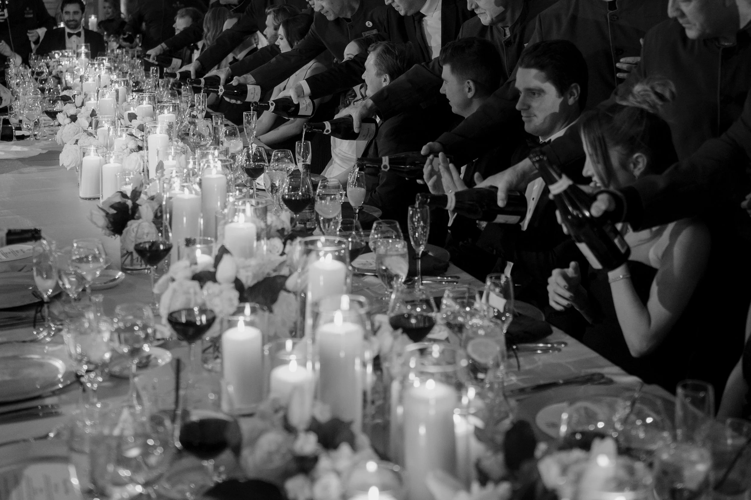 Black and white photo of a formal dinner party with guests in evening attire pouring wine for each other across a long dining table decorated with candles, flowers, and glassware.