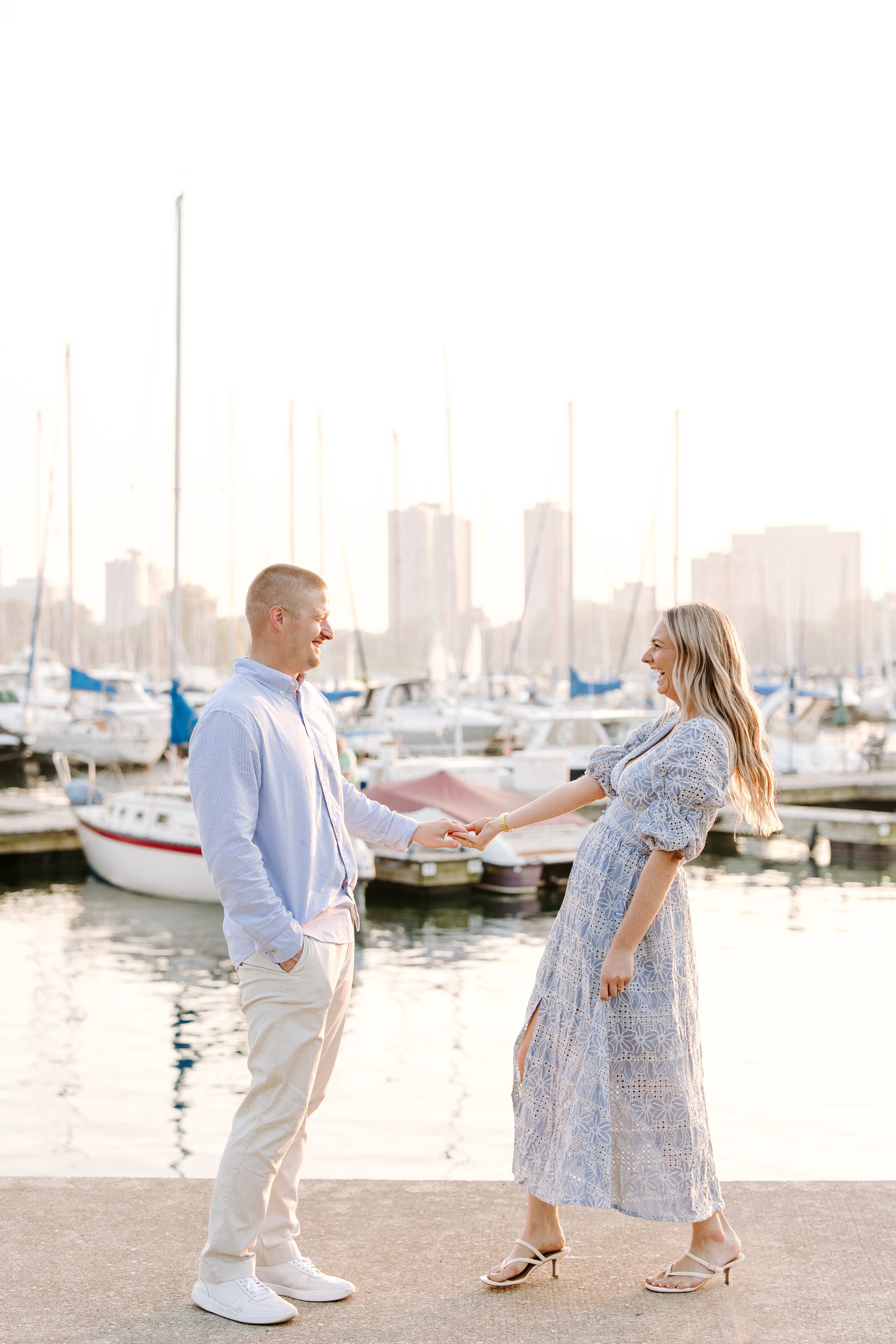 A man and woman standing by a marina, holding hands and smiling at each other with sailboats and high-rise buildings in the background during sunset.