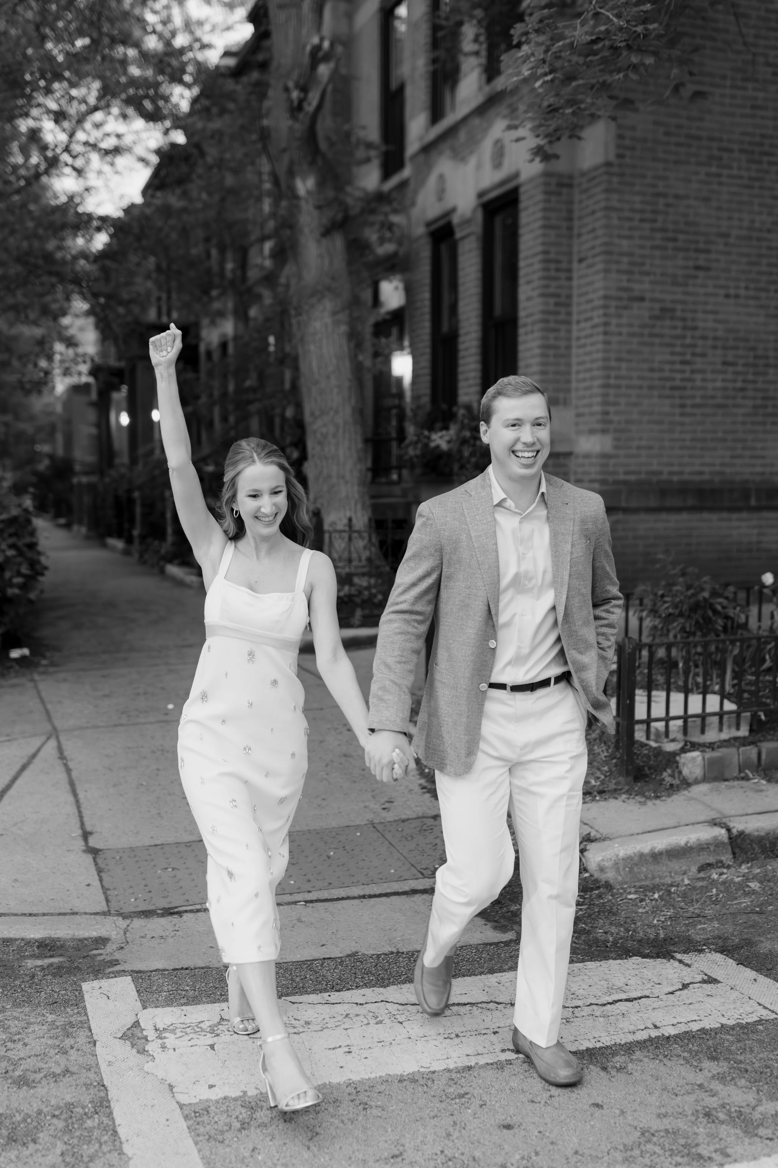 A smiling couple walking hand in hand on the city sidewalk, the woman raising her fist in celebration, in black and white.