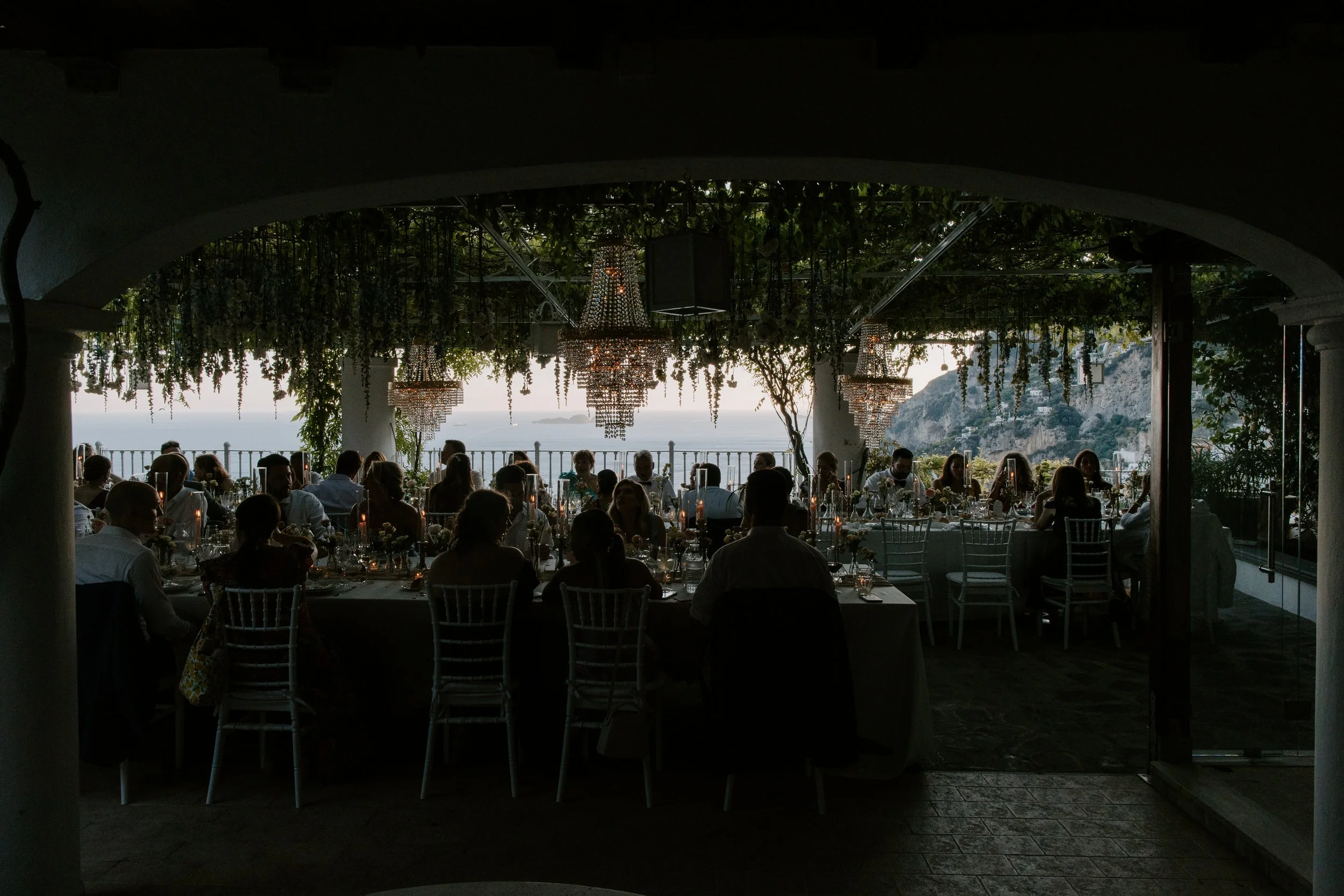 An outdoor restaurant or event space with chandelier lighting, decorated for a gathering or celebration, overlooking a scenic coastal view at sunset with mountainous islands in the distance.