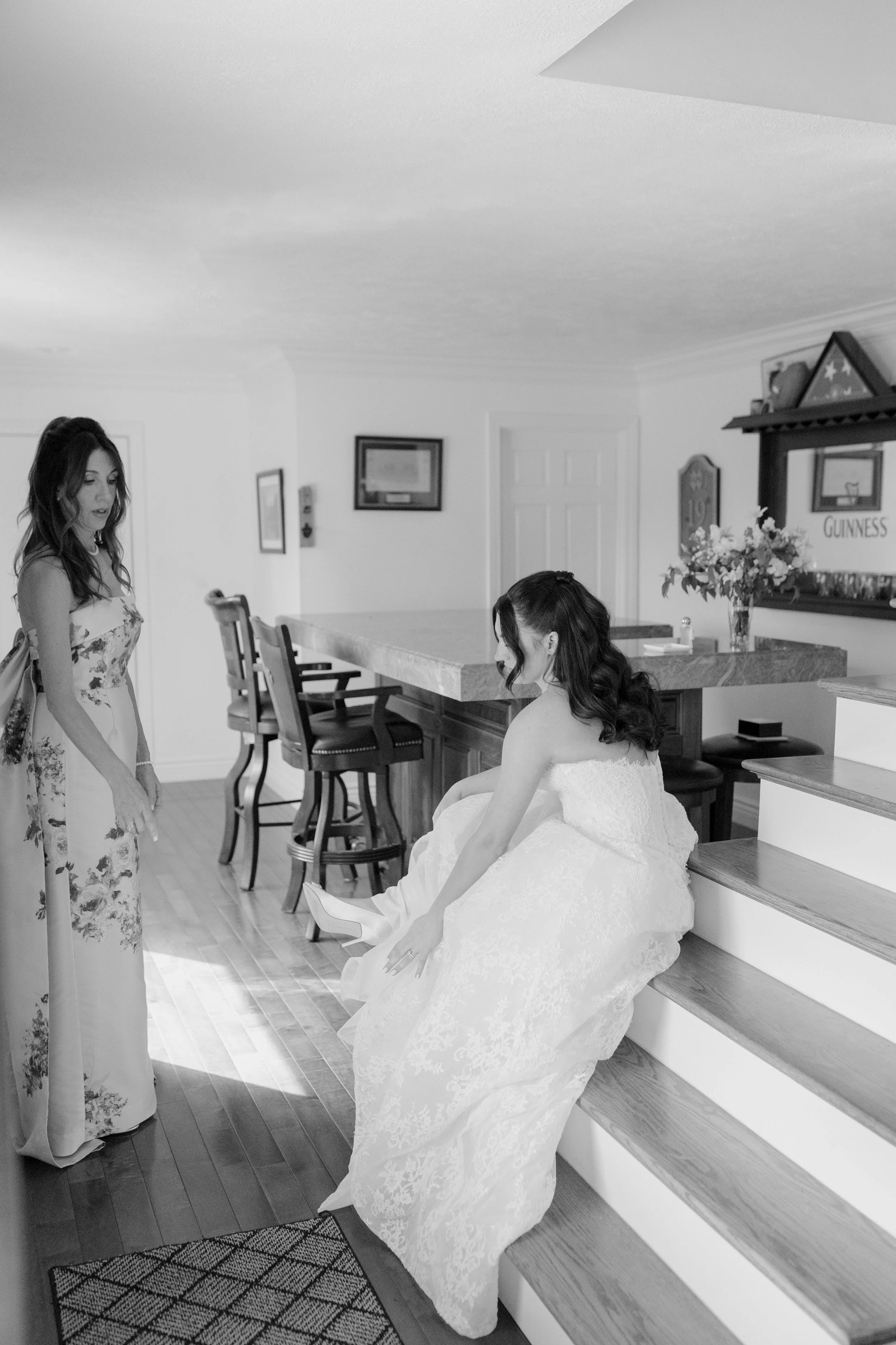 A woman in a wedding gown sitting on stairs and another woman in a floral dress standing nearby in a home setting with a kitchen and decorations in the background.