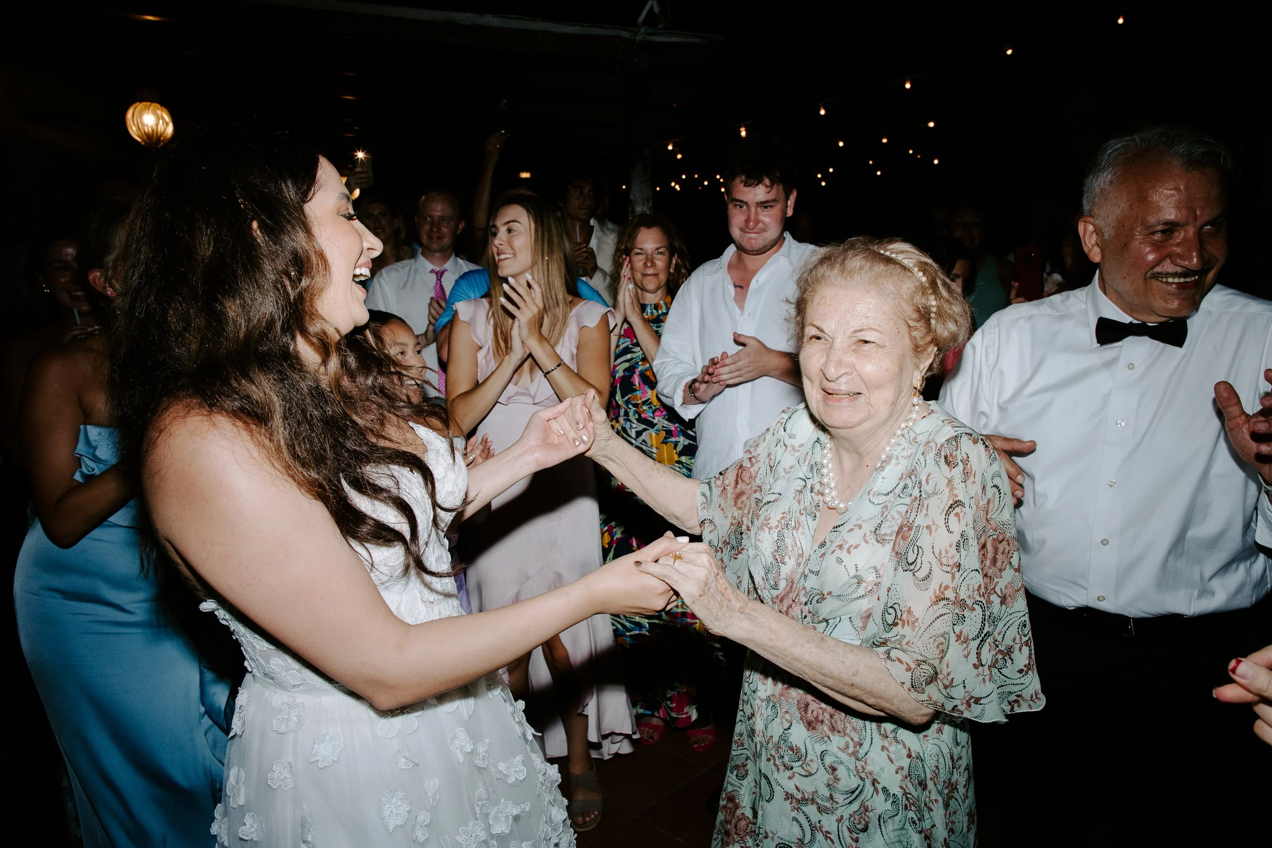 An elderly woman in a floral dress holding hands and dancing with a young woman in a white lace dress at a party. Other guests are seen smiling and clapping in the background, under dim string lights.