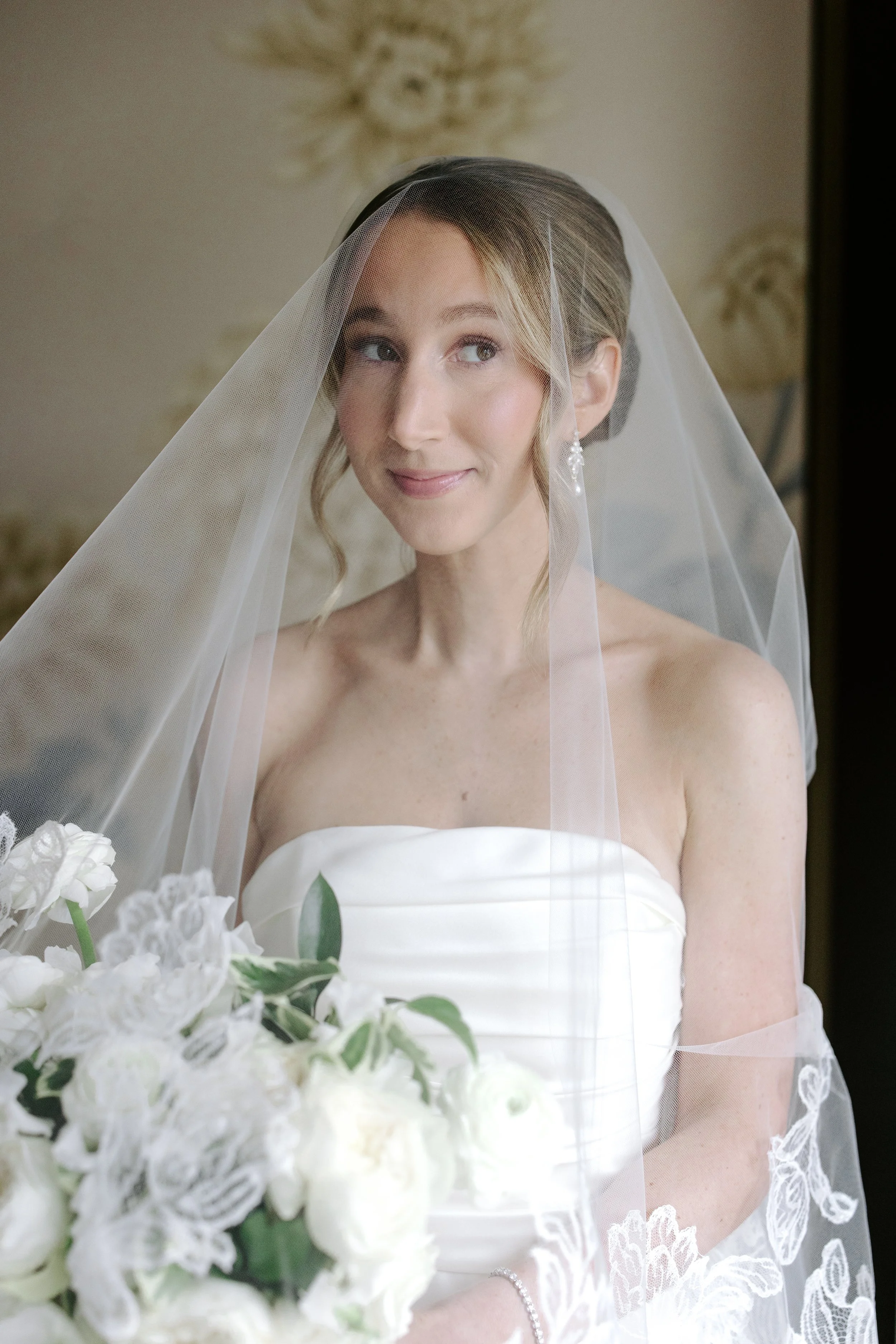 A bride with blonde hair partially covered by a lace veil, wearing a strapless white wedding dress with lace detailing on the bottom, holding a bouquet of white flowers, standing indoors with a beige decorative wall in the background.