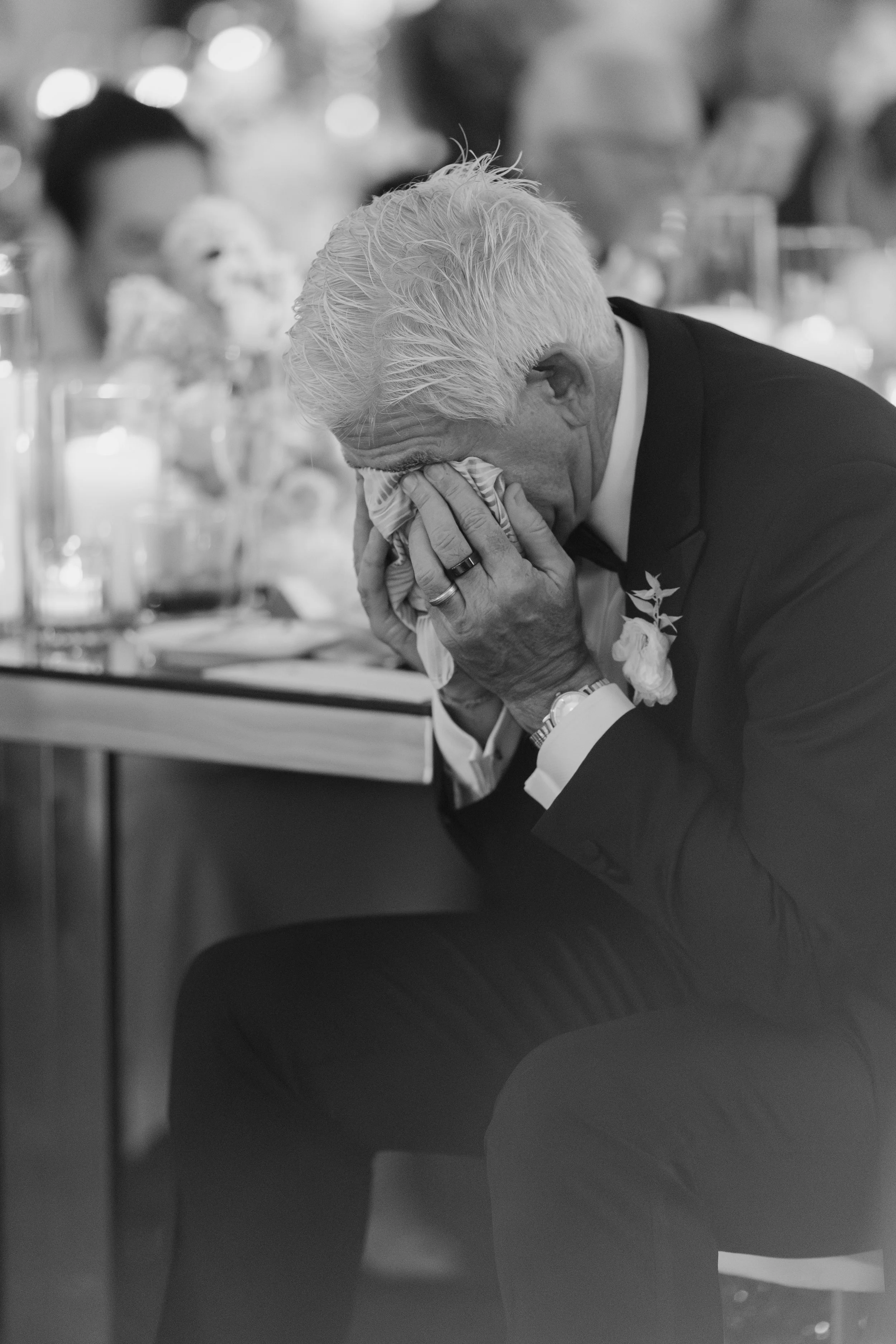 An elderly man with white hair sitting at a table, holding a napkin to his face and appearing emotional, possibly crying, at a formal event.