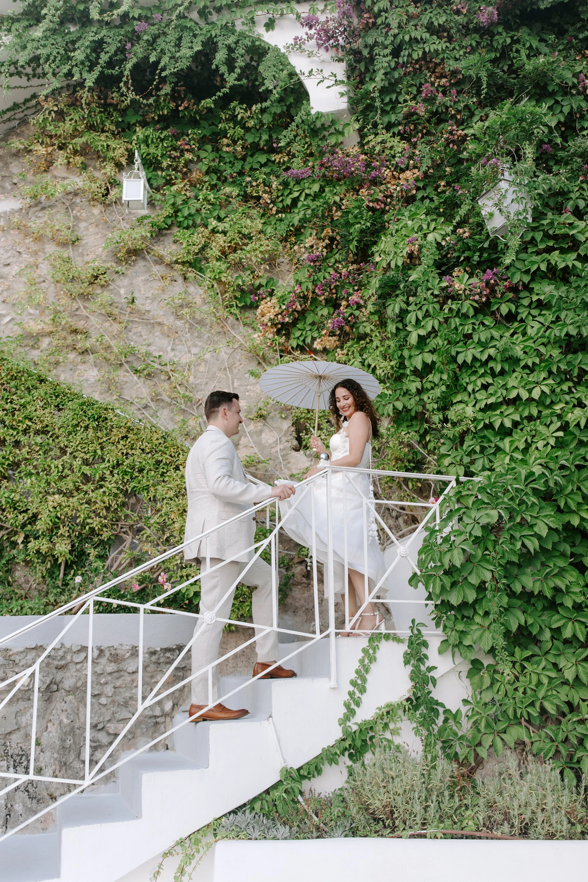 A couple dressed in white wedding attire exchanging vows on a white staircase in front of a green ivy-covered wall, with a woman holding a white parasol.