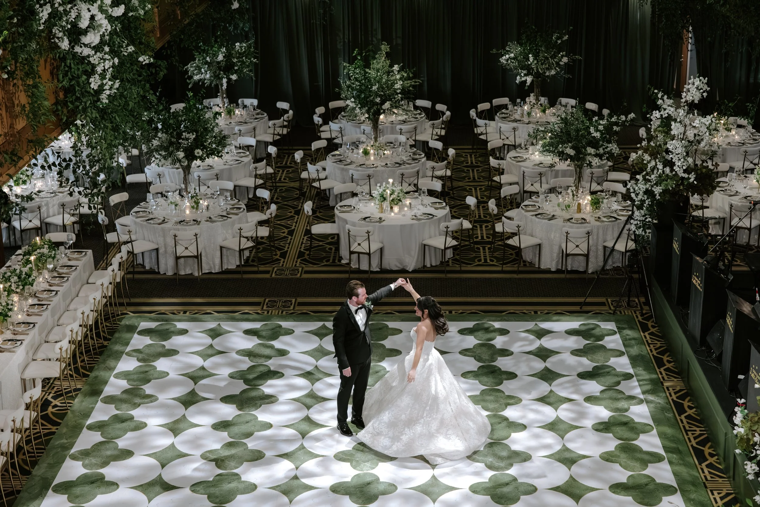 A bride and groom are dancing on a decorated white and green dance floor at a wedding reception with round tables covered in white tablecloths and surrounded by white chairs, with floral centerpieces and candlelit decor, in a banquet hall with dark c