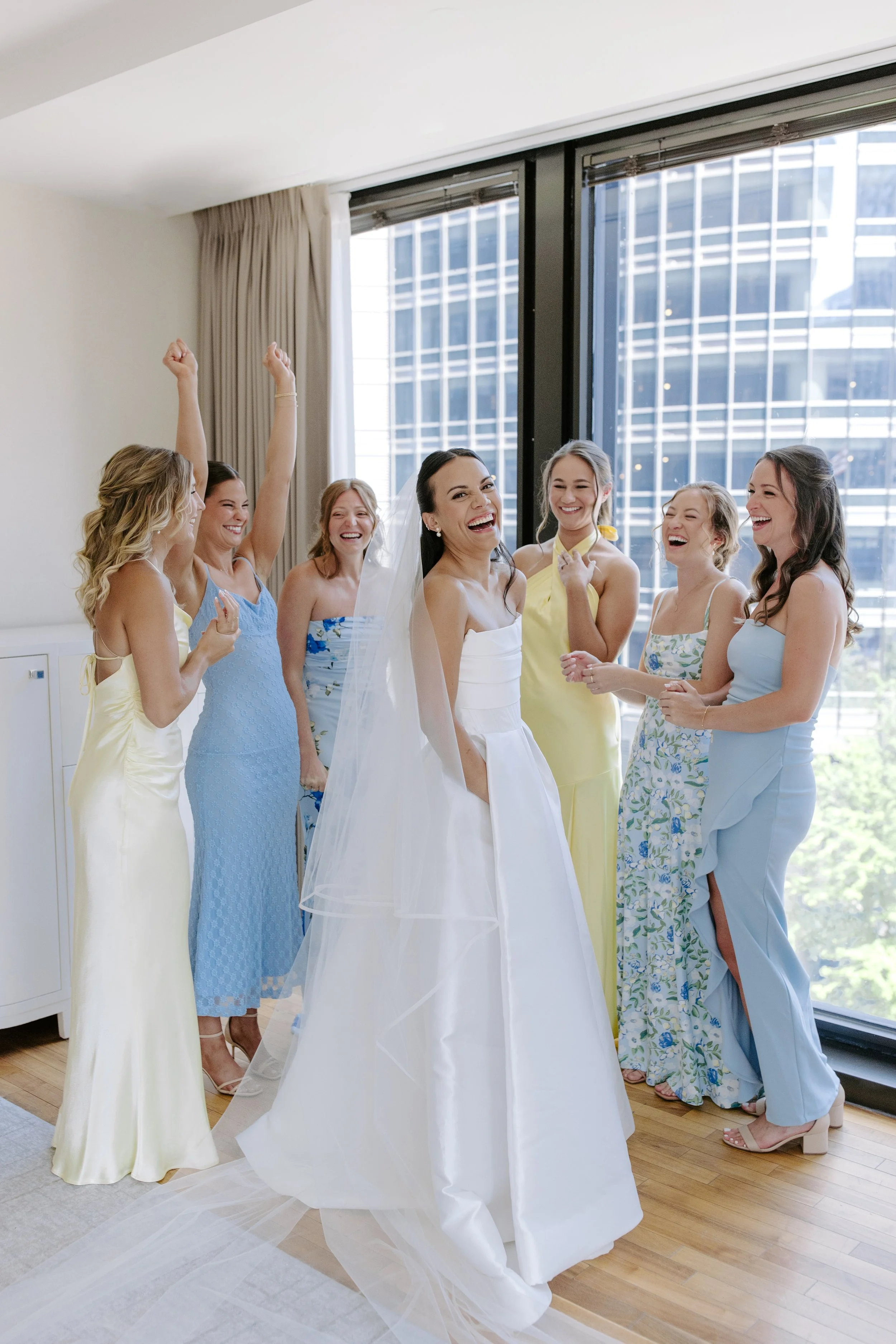 Bride in a white wedding gown smiling with a group of women in colorful dresses at a wedding celebration.