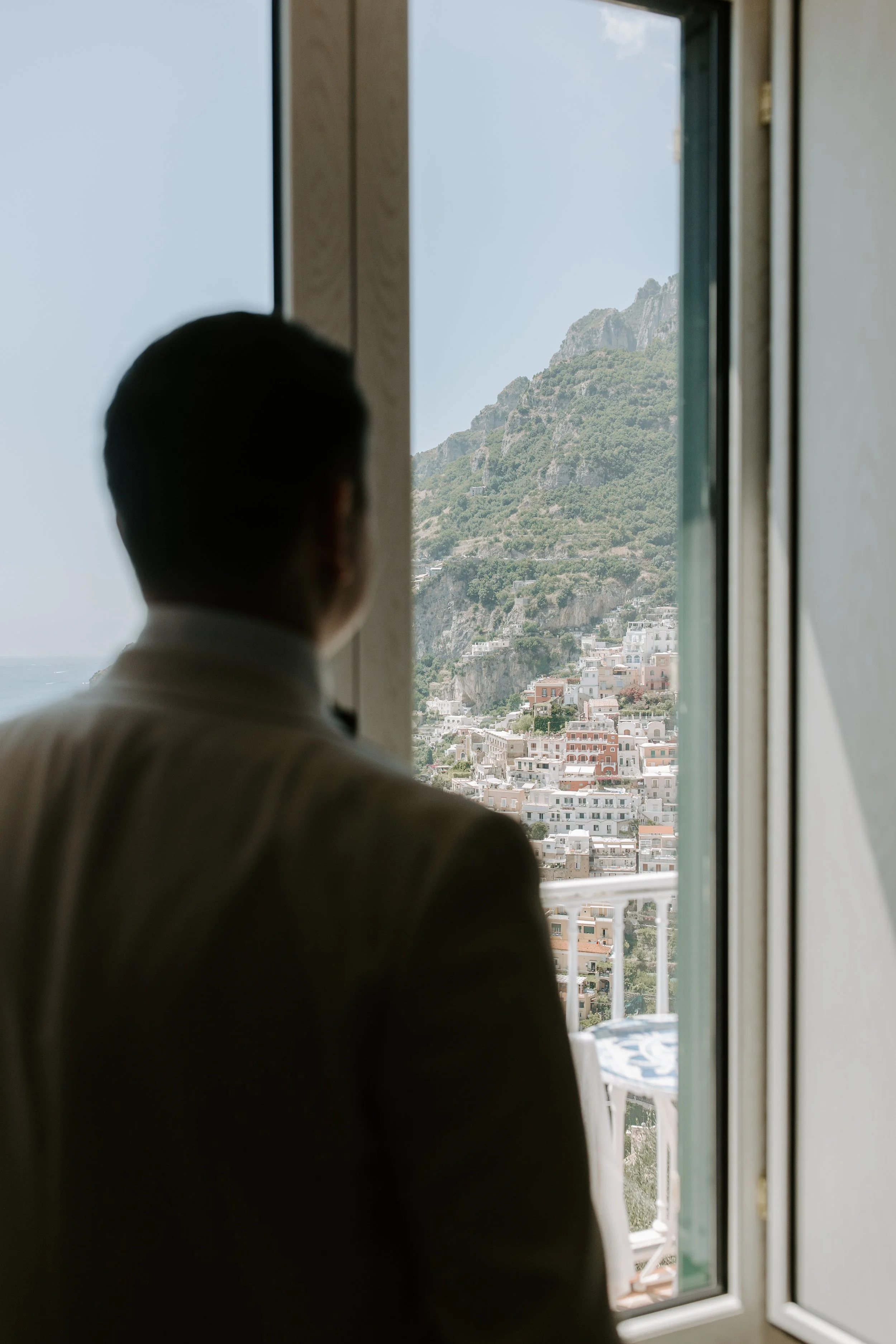 A man in a beige suit stands on a balcony, gazing at a mountain with green vegetation and a town below, framed by an open window.