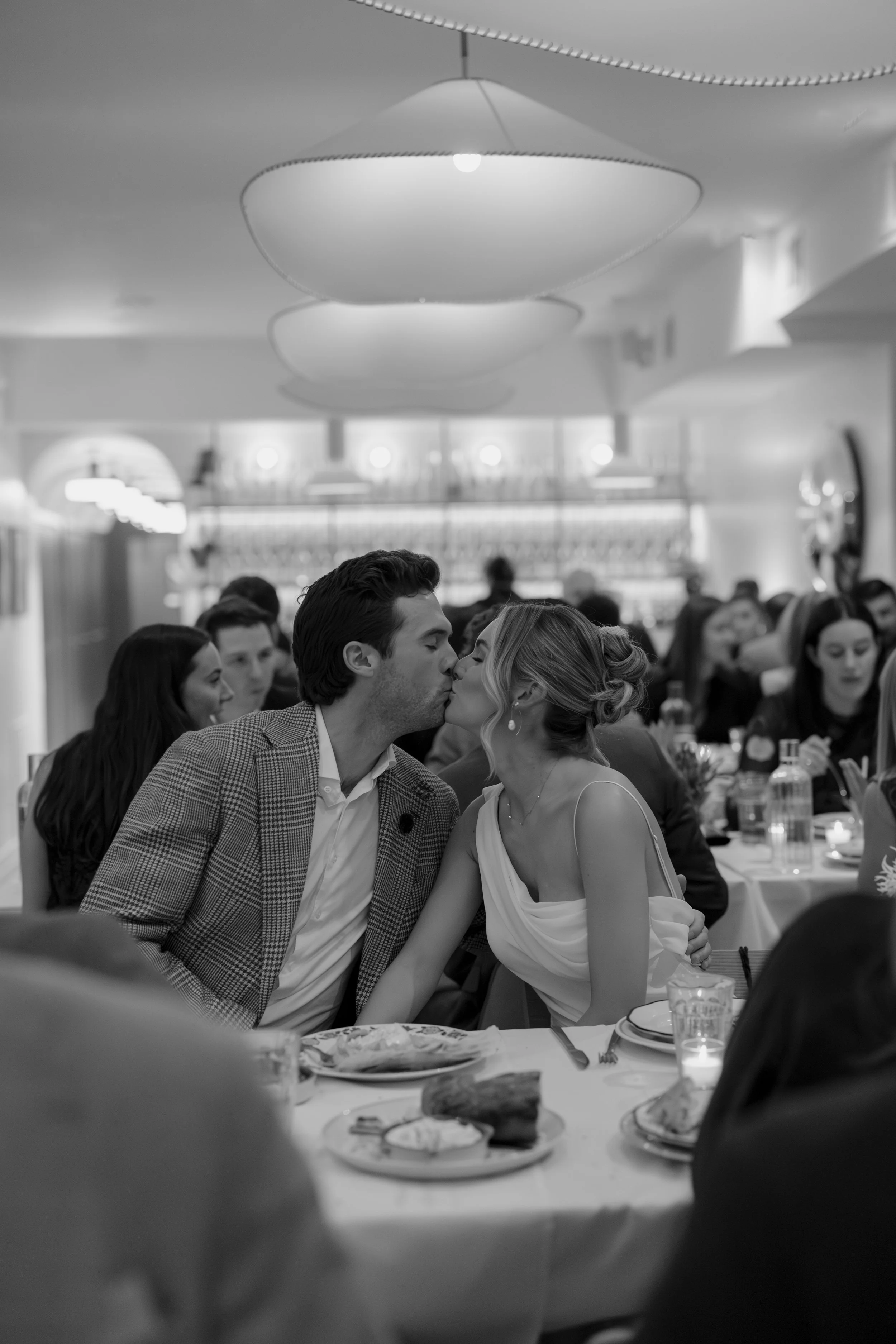 A couple at a wedding reception is kissing. The man has dark hair and is wearing a checked suit, and the woman has blonde hair and is dressed in a white gown. They are seated at a table with food and drinks, surrounded by other guests in a well-lit v