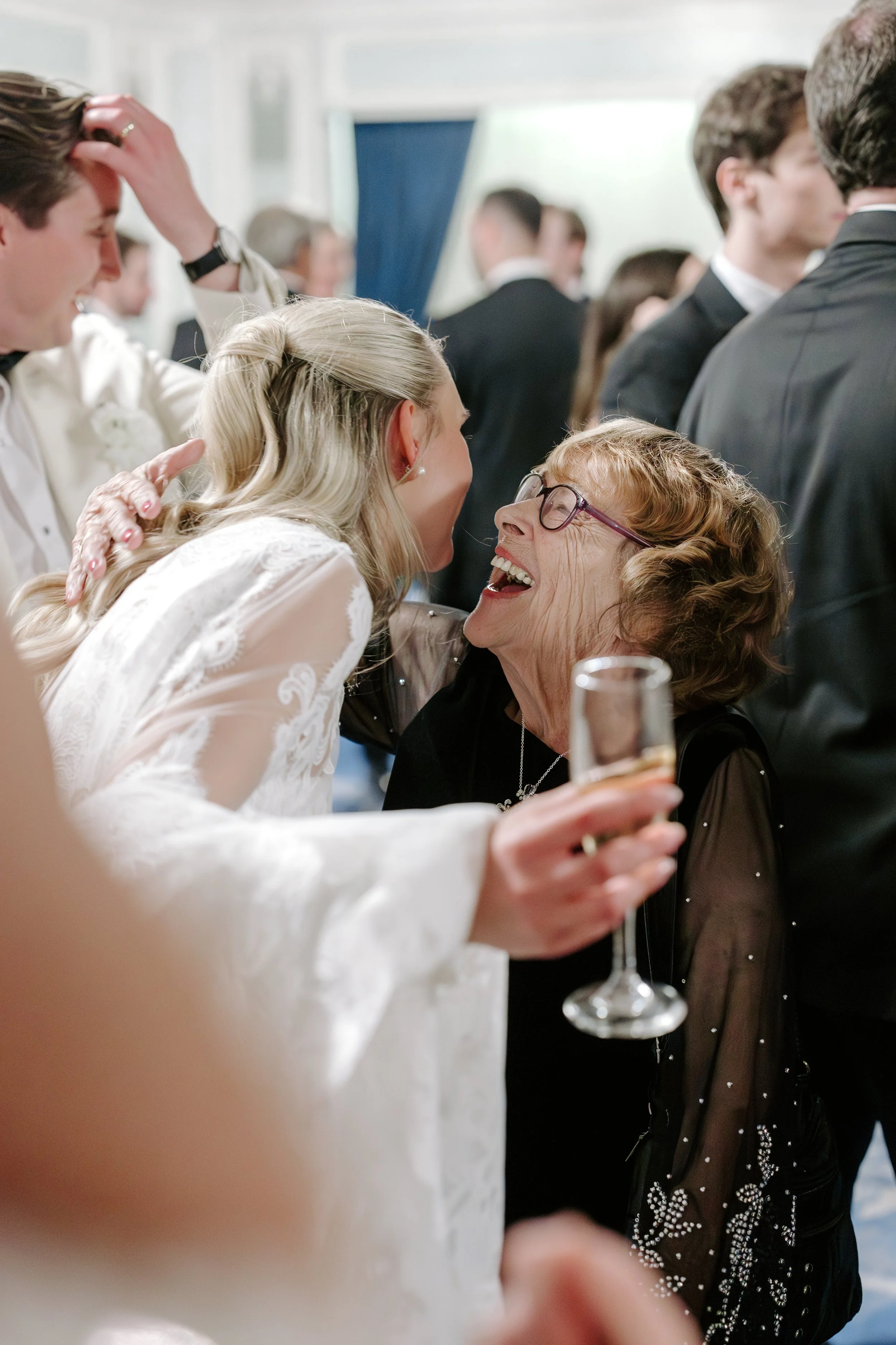 A joyful bride and an older woman with glasses sharing a hug at a wedding reception, with guests in formal attire in the background.