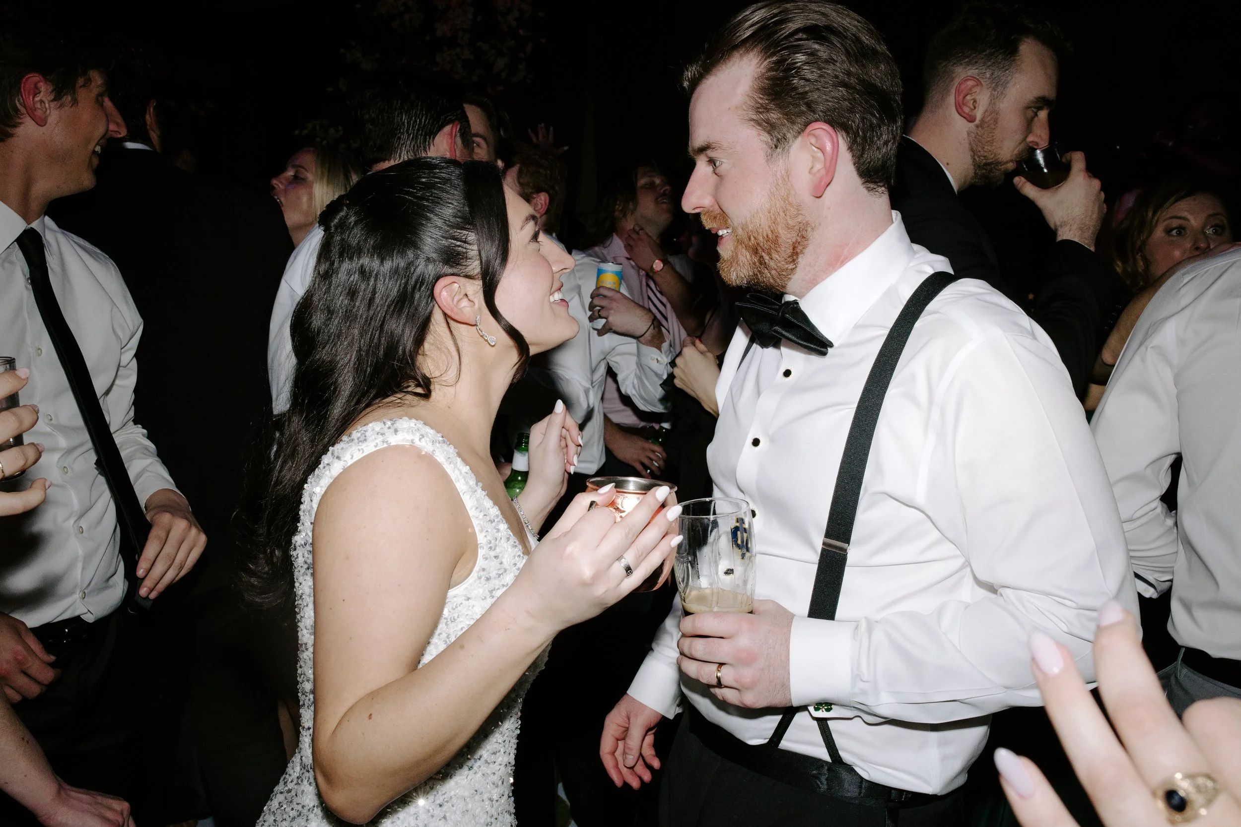 A crowded party scene with a woman in a white dress smiling and holding a drink, talking to a man with a beard in a white shirt and black tuxedo, surrounded by other people enjoying drinks and conversation.