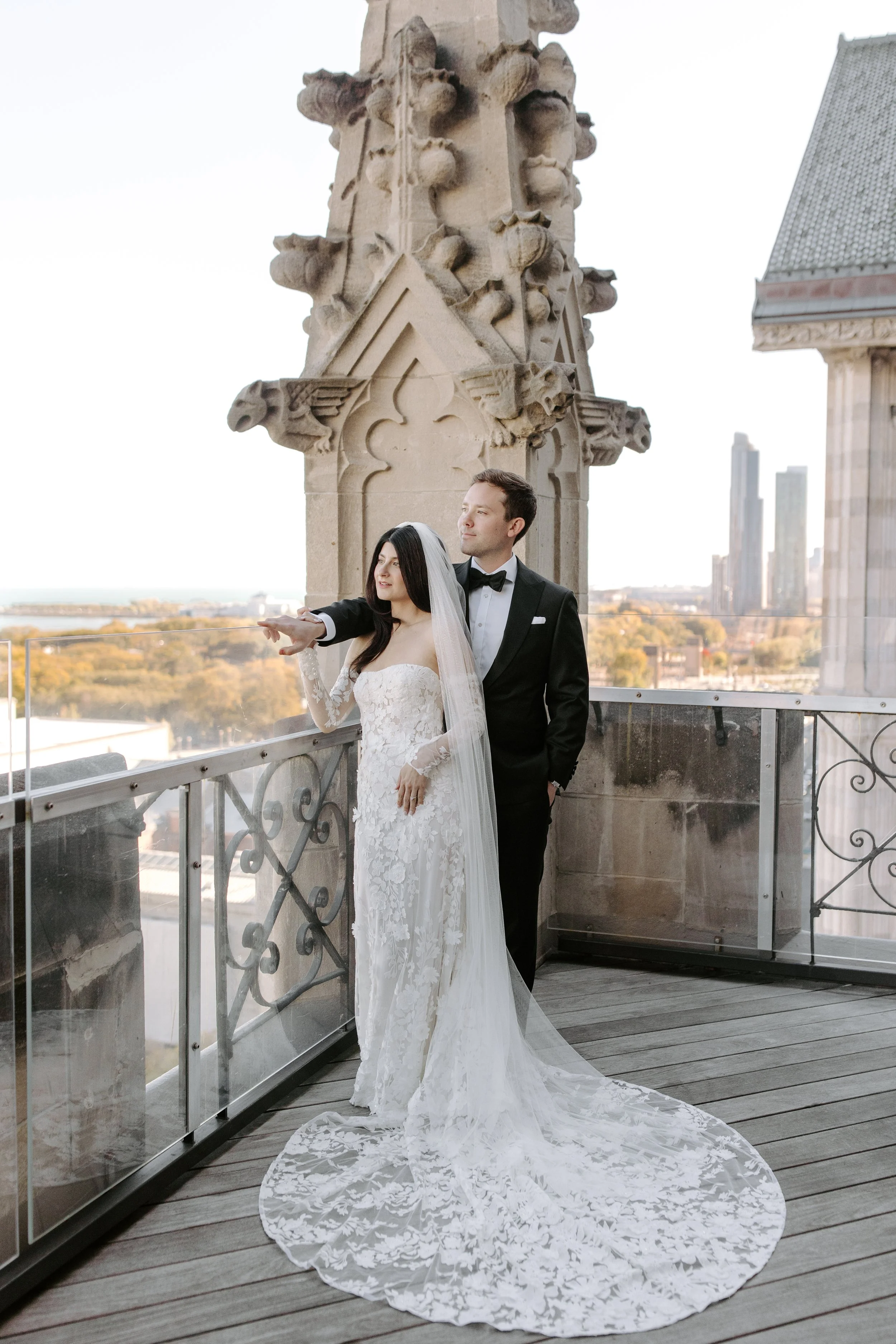 A bride and groom standing on a balcony with ornate metal railing, in front of a stone Gothic-style architectural feature with intricate carvings, overlooking a cityscape with tall buildings and trees in the distance. The bride is dressed in a white 