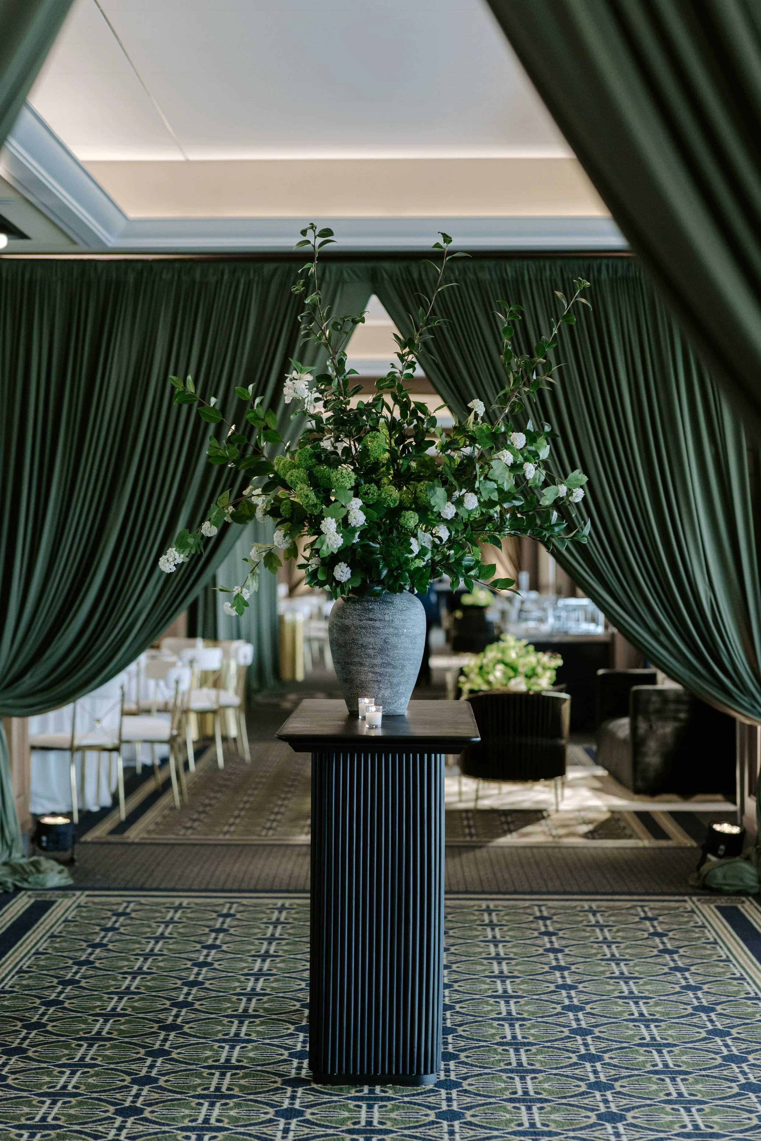 Interior space with dark green curtains framing a floral arrangement in a gray vase on a black pedestal table, with a patterned carpet, chairs, and tables in the background.
