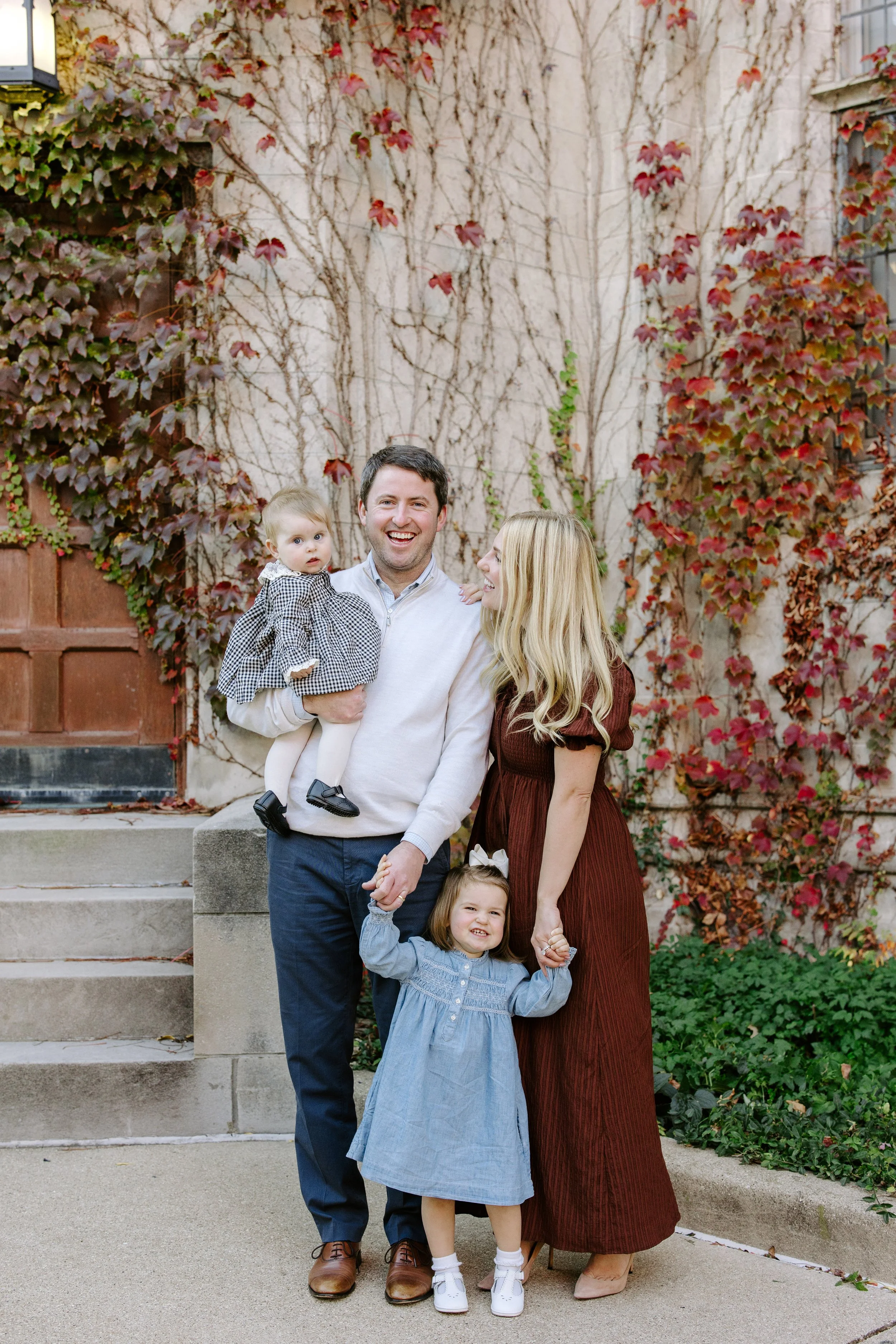 Family of five standing outdoors on steps, smiling and holding hands, with a brick wall and colorful autumn ivy in background.
