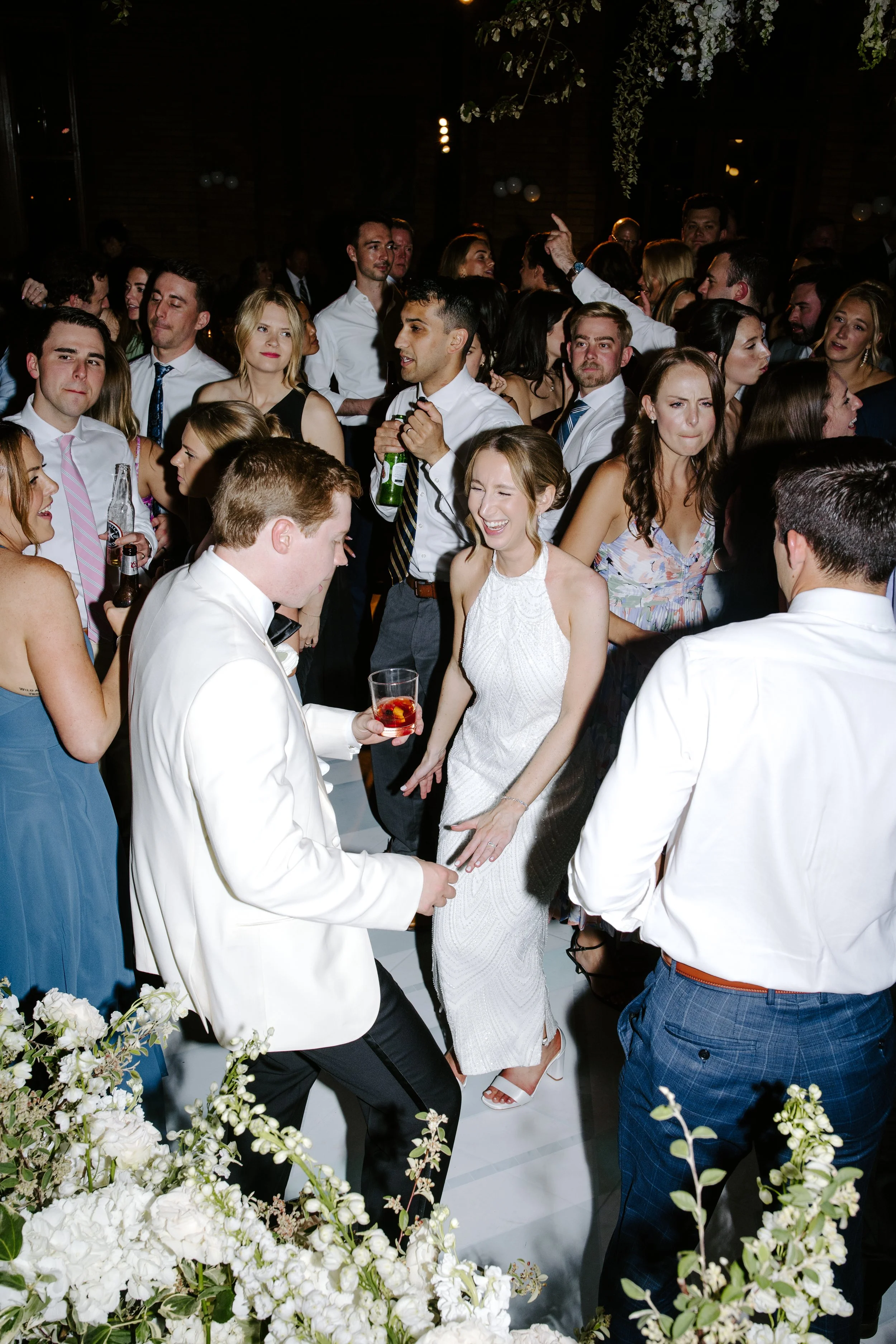 People dancing and socializing at a lively wedding reception or party, with floral decorations in the foreground and dim lighting in the background.