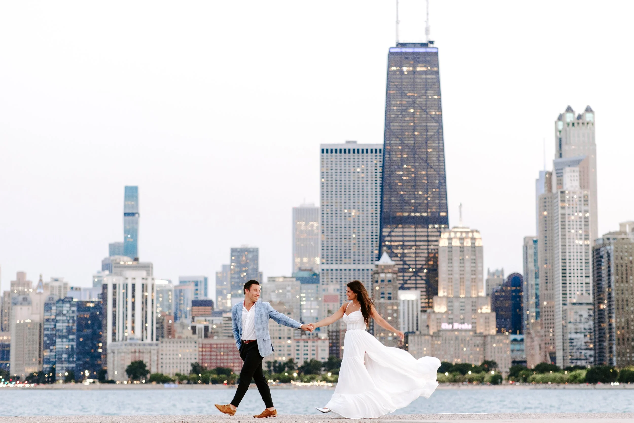 A couple dancing by a body of water with a city skyline, including the One World Trade Center, in the background at dusk.