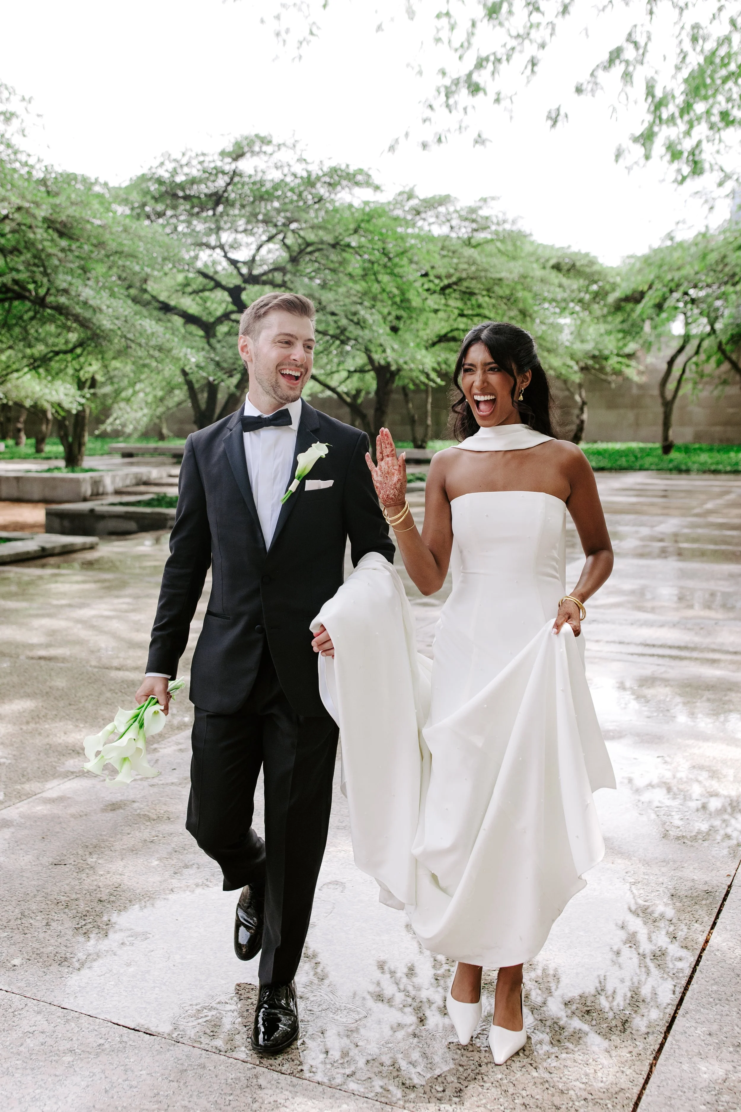 A joyful bride and groom walking outdoors on a rainy day, with the bride wearing a white strapless wedding gown and white high heels, and the groom dressed in a black tuxedo with a bow tie, holding a small bouquet of white flowers. They are smiling and celebrating, with lush green trees in the background.
