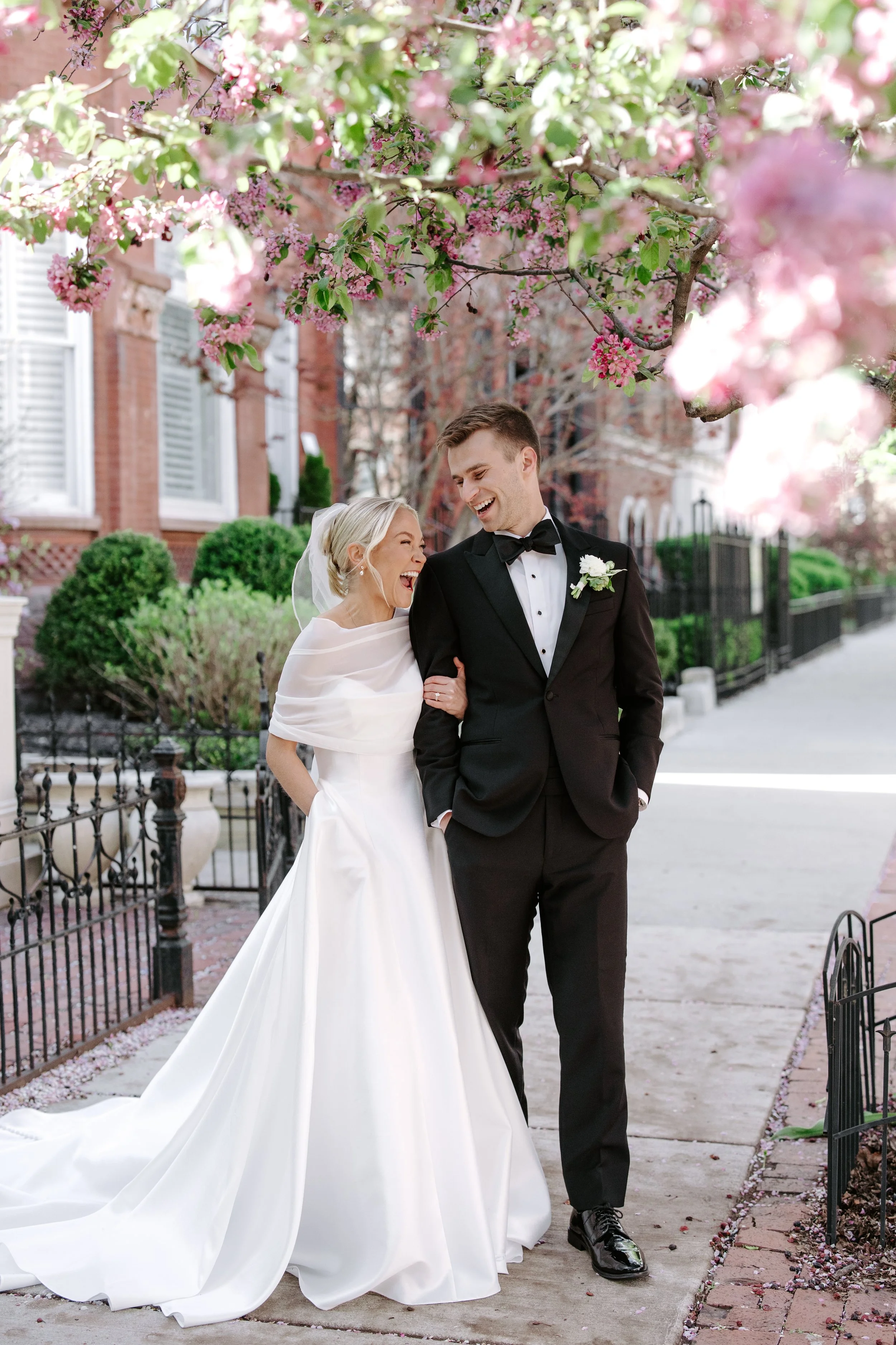 A bride and groom walking outdoors under pink flowering trees, smiling and laughing. The bride wears a white wedding dress with a long train and veil, while the groom wears a black tuxedo with a bow tie and boutonniere. They are on a sidewalk with a 