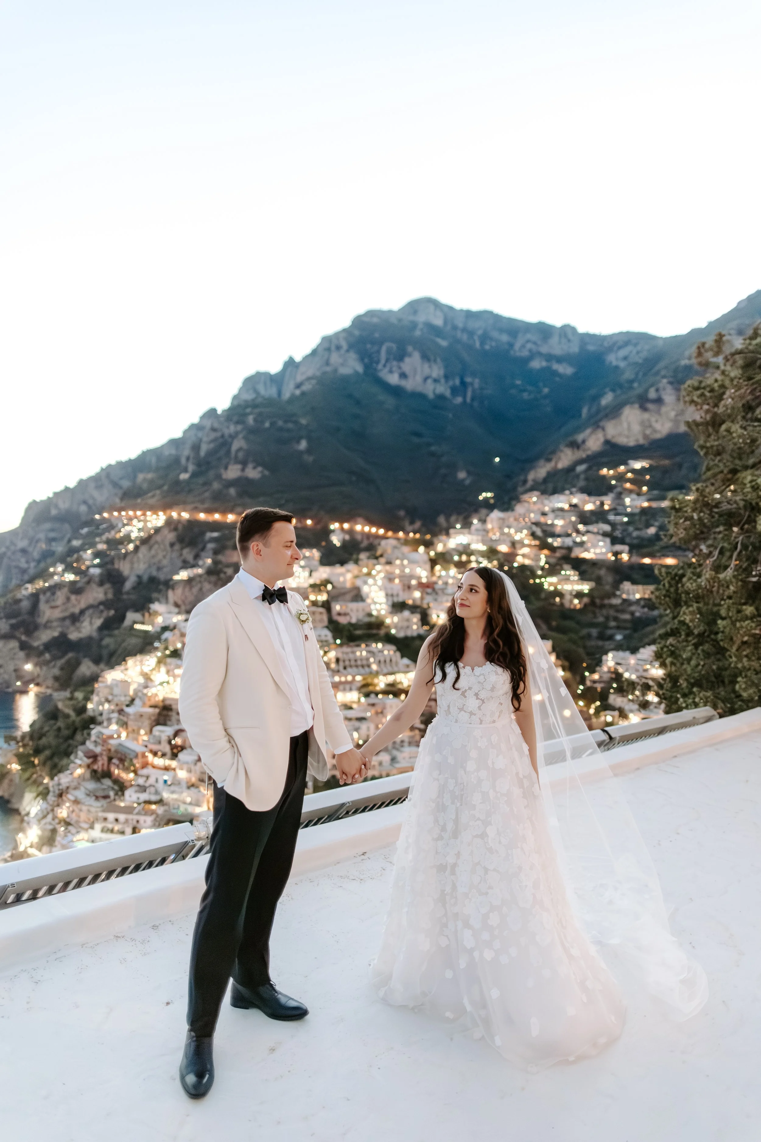 A bride and groom holding hands, standing on a balcony overlooking a hillside town illuminated with lights at dusk.