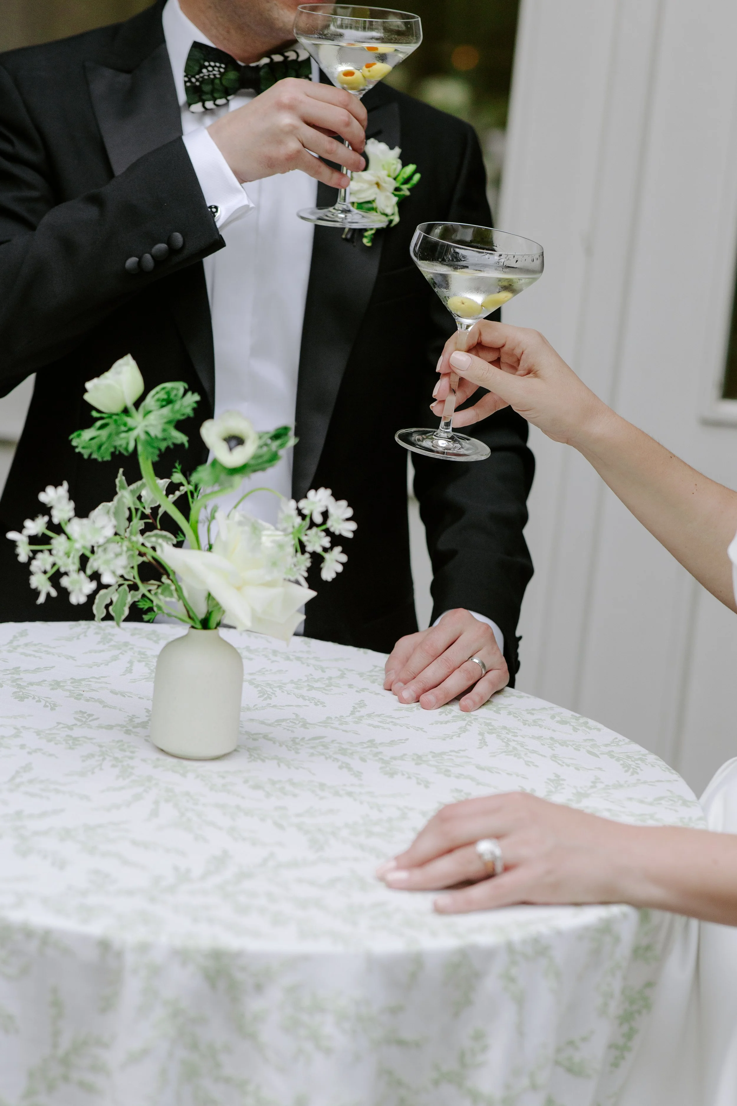 A man in a tuxedo holding a martini glass with olives, and a woman holding another martini glass with olives, sitting at a table with a floral centerpiece in a white vase.