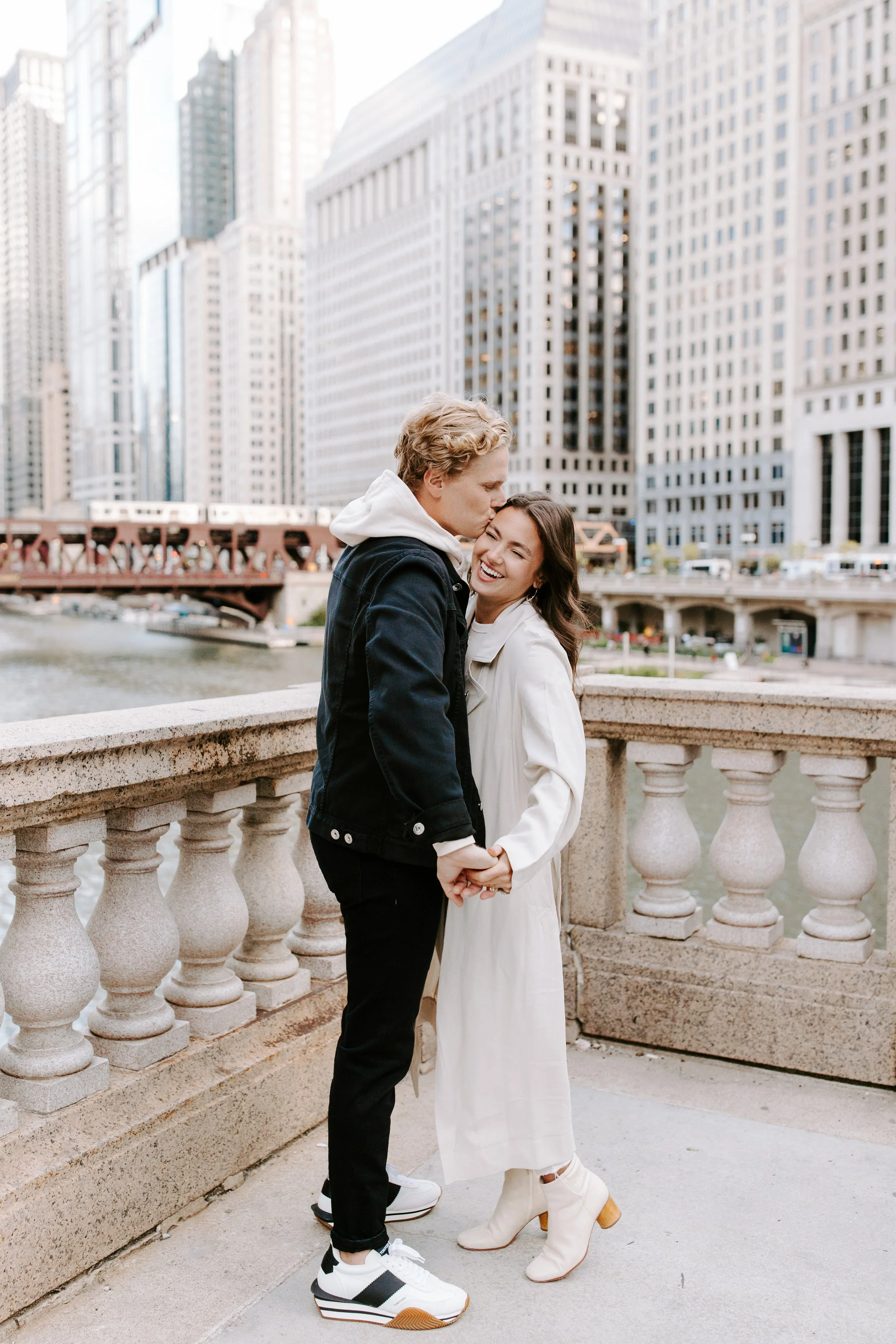 A couple standing on a stone bridge in front of a city river, with tall skyscrapers in the background. The man is kissing the woman's forehead, and they are holding hands. The woman is wearing a white dress and tan high-heeled boots; the man is dress