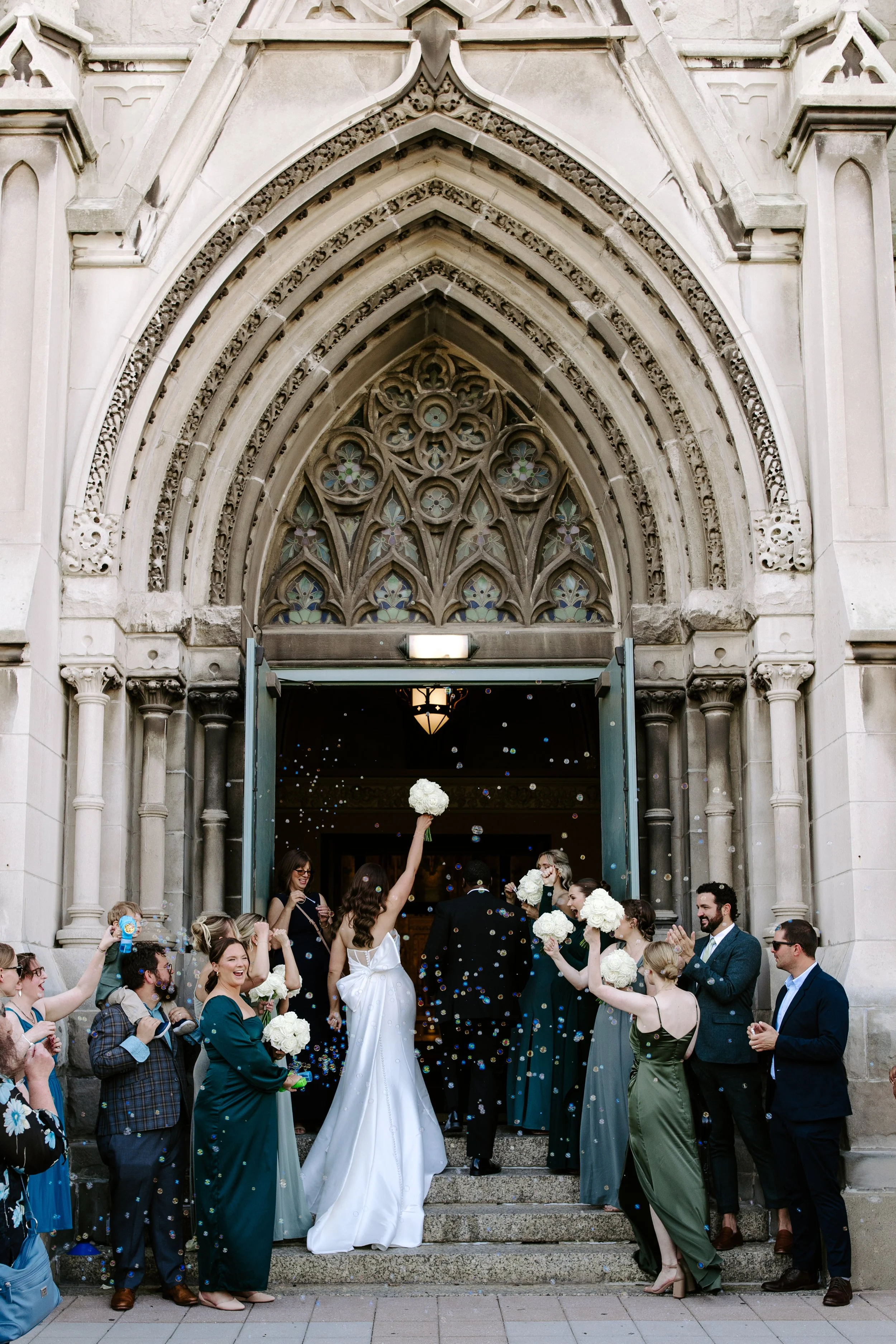 A group of people celebrating outside a church during a wedding reception, with some guests throwing confetti and holding bouquets of white flowers. The bride and groom are at the center, surrounded by friends and family.