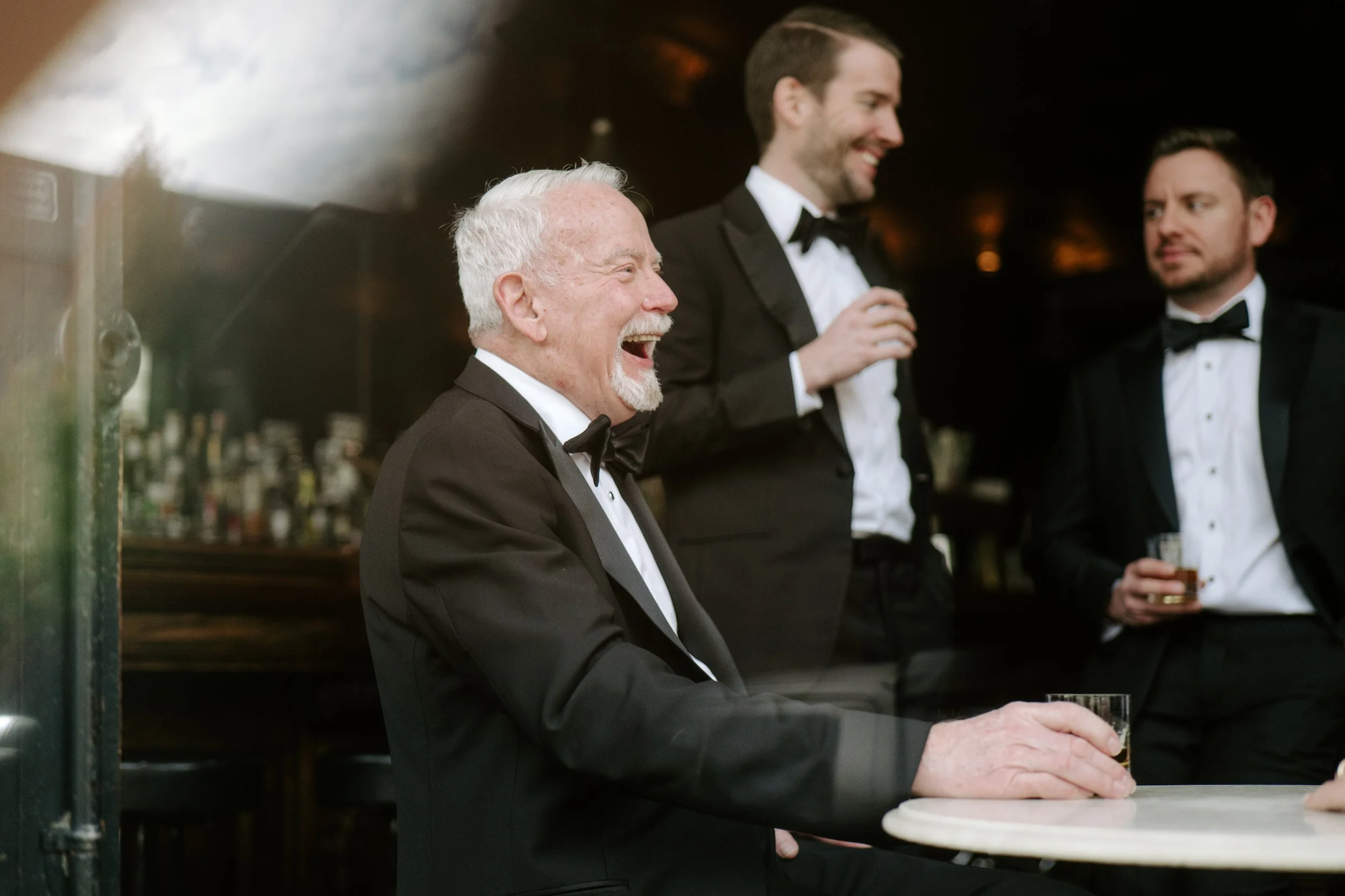 A white-haired man in a tuxedo laughing and holding a drink, standing at the bar with two other men in tuxedos, one with a beard and the other with glasses, inside a dimly lit bar or lounge.