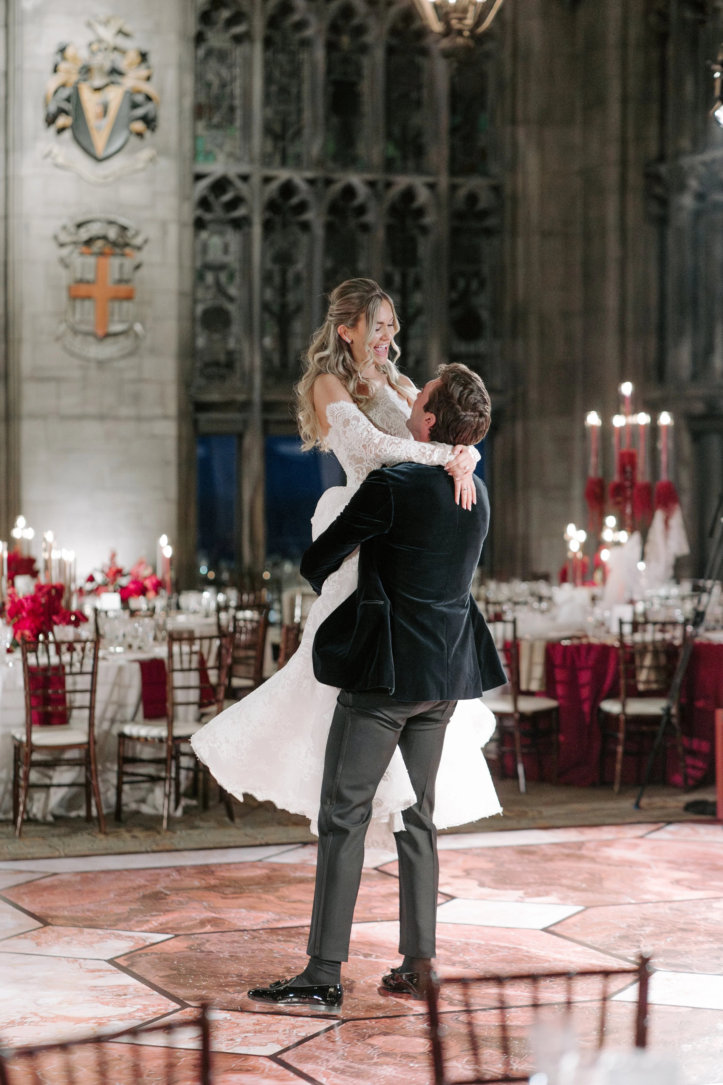 A bride and groom dancing in a decorated church reception hall with tables and chairs, floral arrangements, and candles in the background.