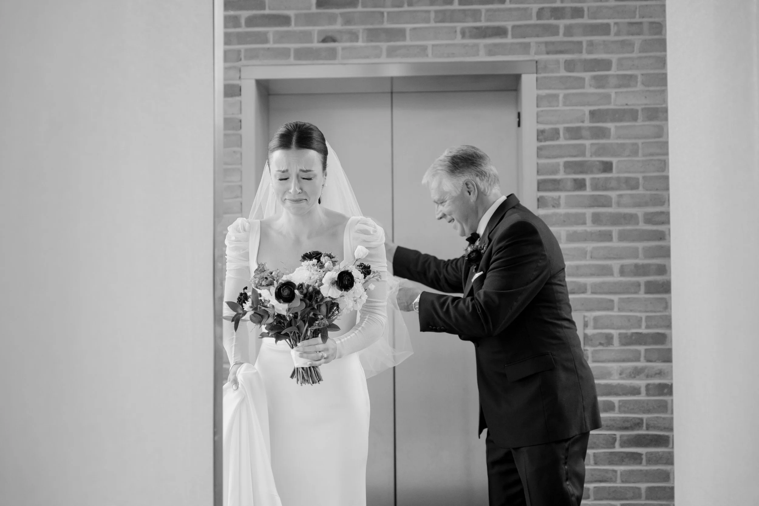 Black and white photo of a bride with a sad expression holding a bouquet of flowers, and a man in a suit smiling and looking down, standing against a brick wall.