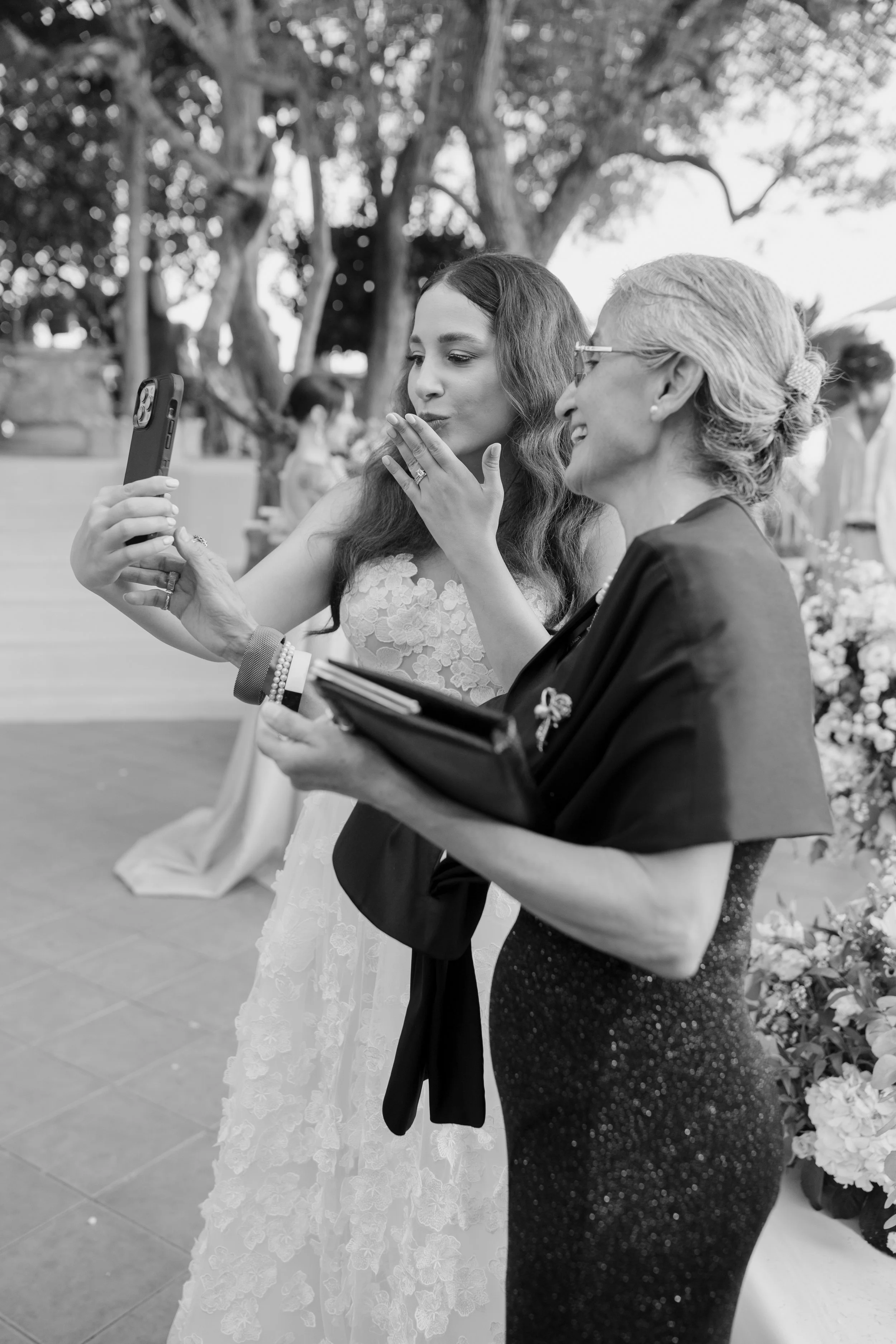Two women are looking at a smartphone together. One woman, wearing a white dress with lace details, appears to be blowing a kiss or making a gesture, while the other woman, dressed in dark attire and glasses, is smiling and holding a folder. They are