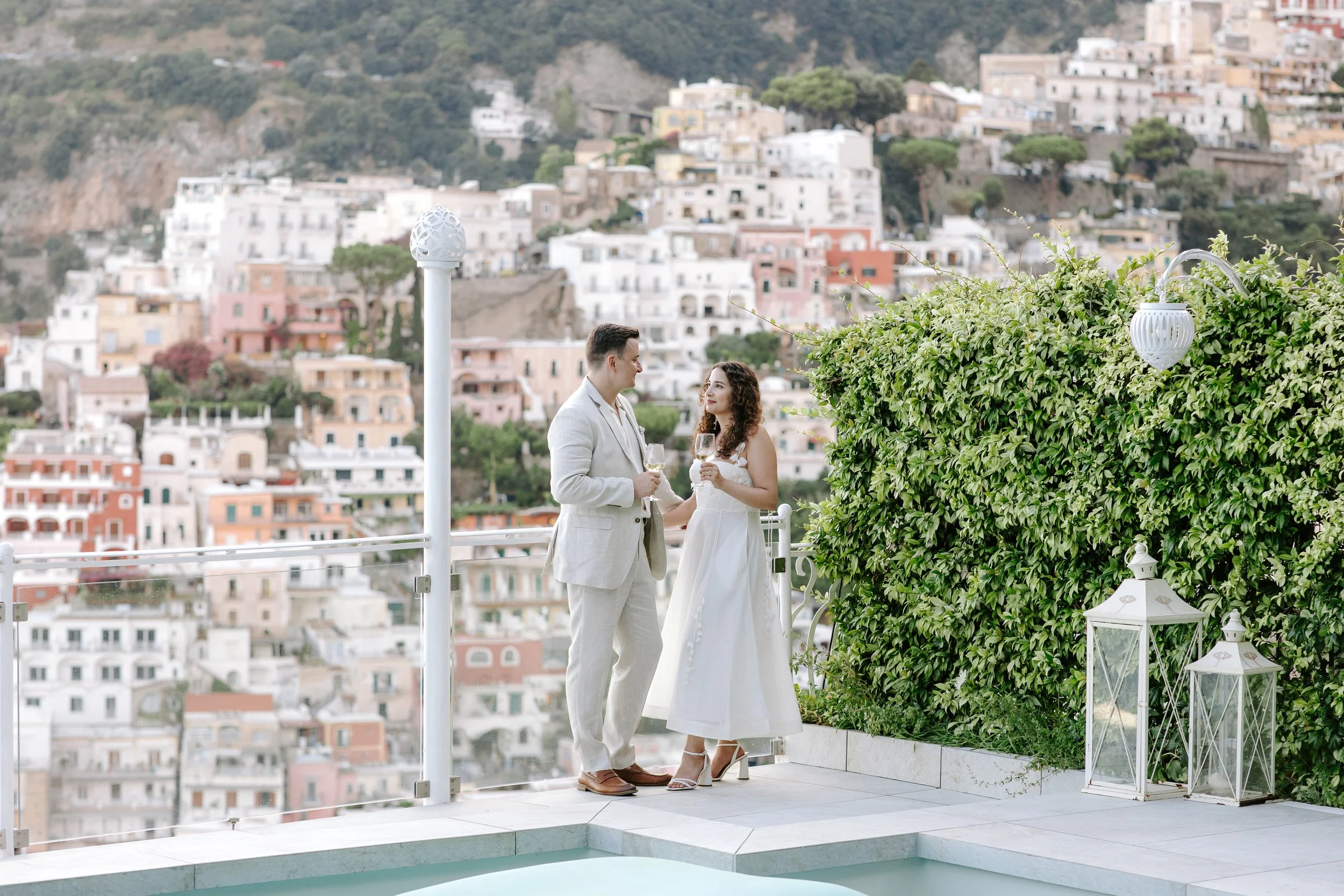 A couple dressed in white on a balcony overlooking a hillside town, holding wine glasses and gazing at each other.