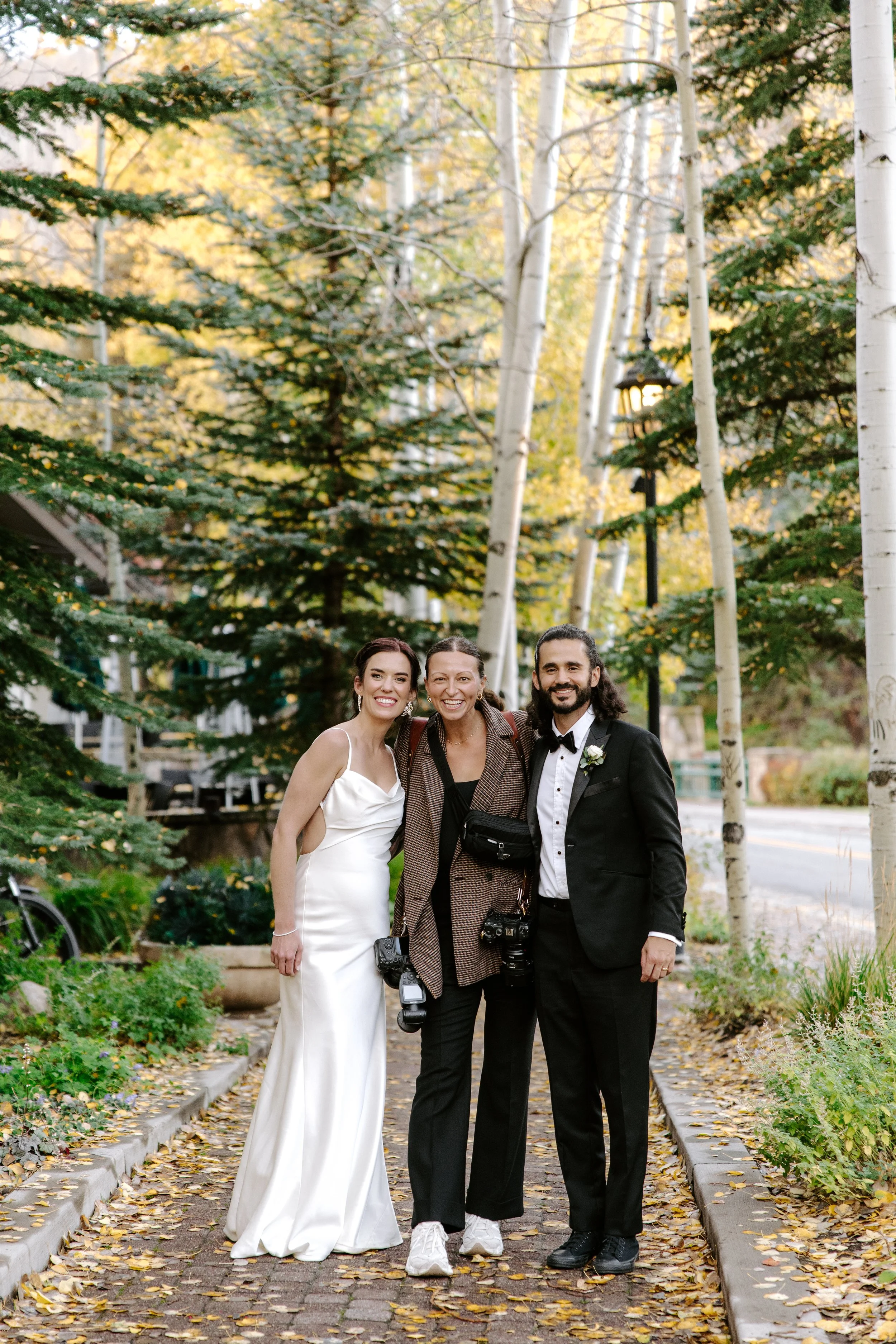 Three people standing together on a tree-lined cobblestone path during fall, smiling at the camera. One woman in a white wedding dress, a woman in casual clothing, and a man in a tuxedo with a boutonniere.
