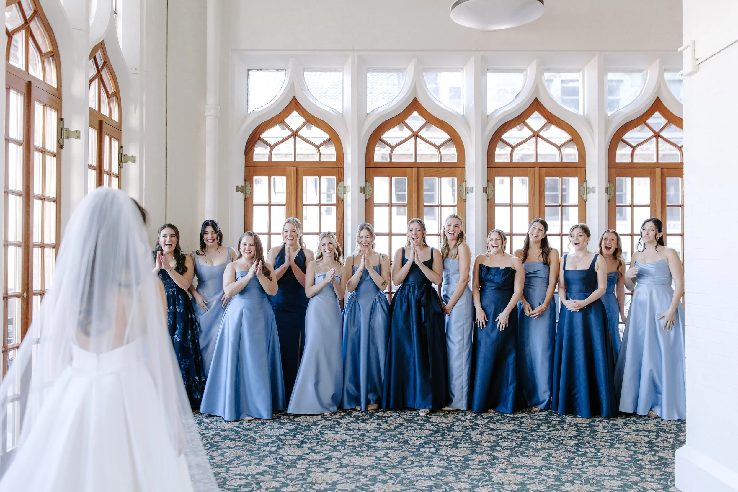 A bride in a white wedding dress with her back to the camera, facing a group of women dressed in matching blue dresses, inside a bright room with large Gothic-style windows.