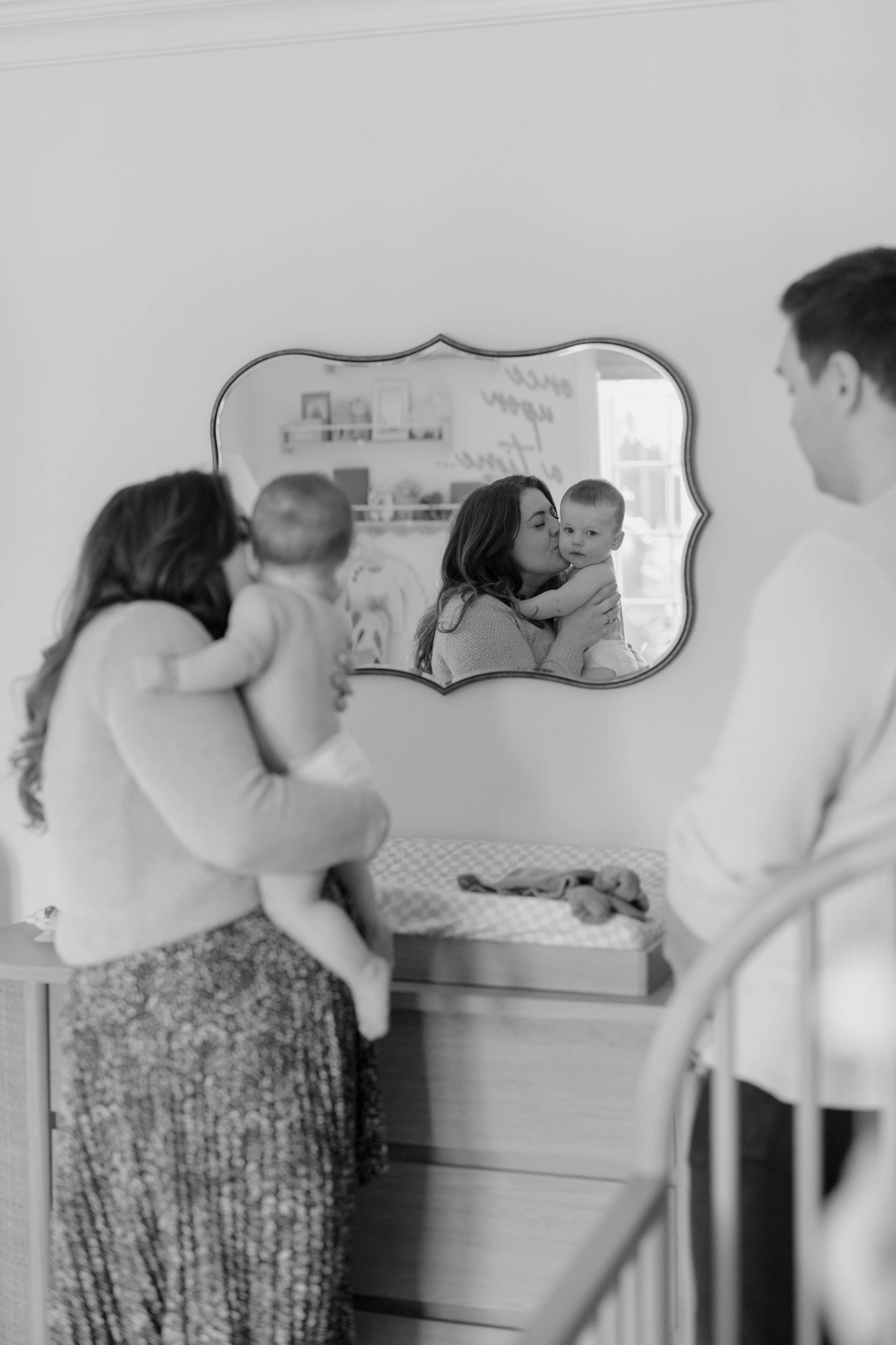 A black-and-white photo of a family of four in a cozy room. The mother is holding and kissing her young child in front of a mirror. The father and another woman are nearby, with the woman holding a baby. The mirror reflects the scene, showing the mot