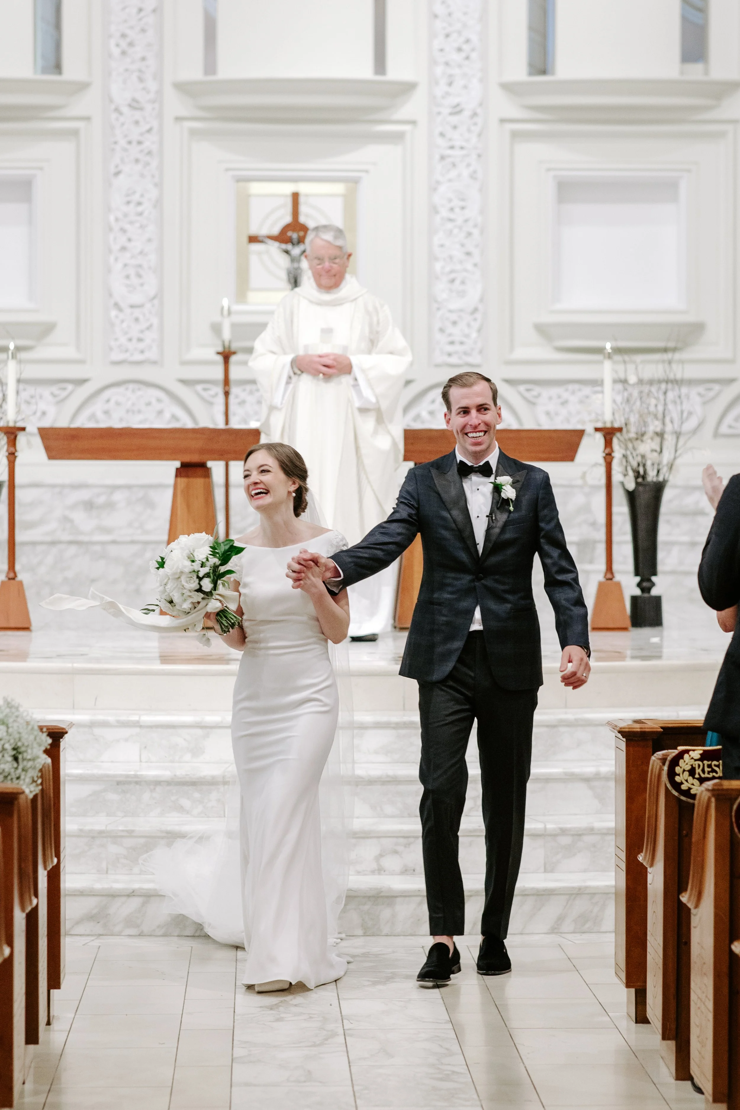 A newlywed couple walking down the aisle of a church after their wedding ceremony. The bride is holding a bouquet of white flowers, and the groom is smiling and holding her hand. The officiant is standing behind them, a priest in white robes. The chu
