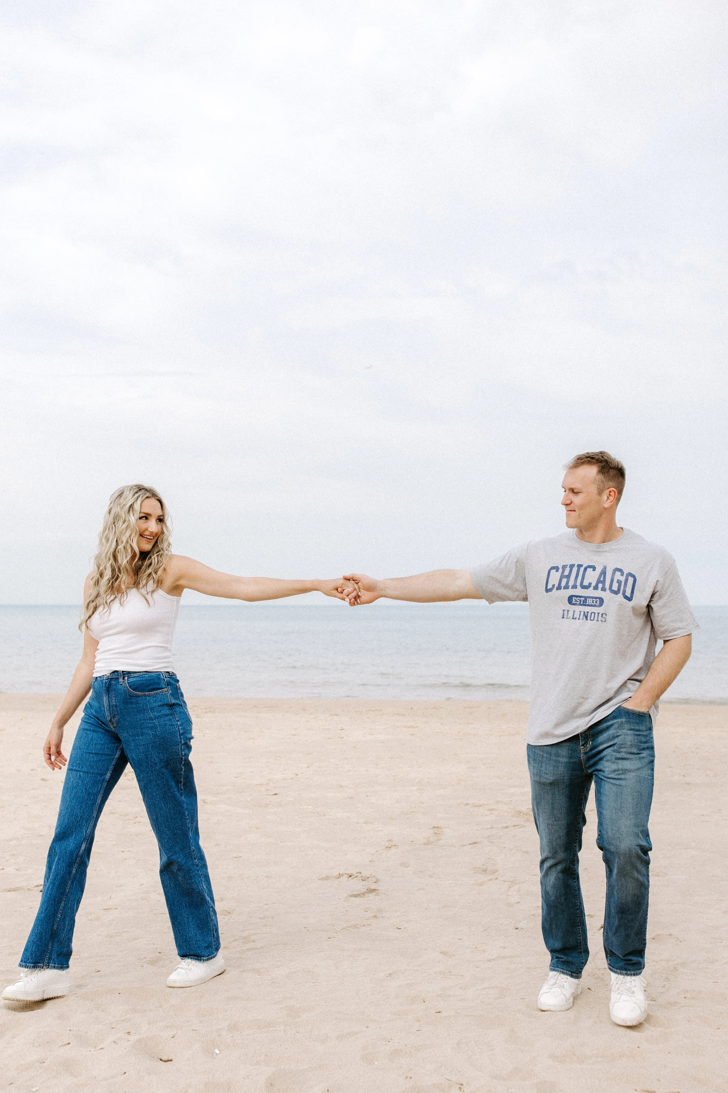 A young woman and a young man standing on a sandy beach, holding hands across his chest. The woman is smiling and wearing a white tank top and blue jeans, the man is wearing a gray "Chicago Illinois" t-shirt and blue jeans, and they are both looking 