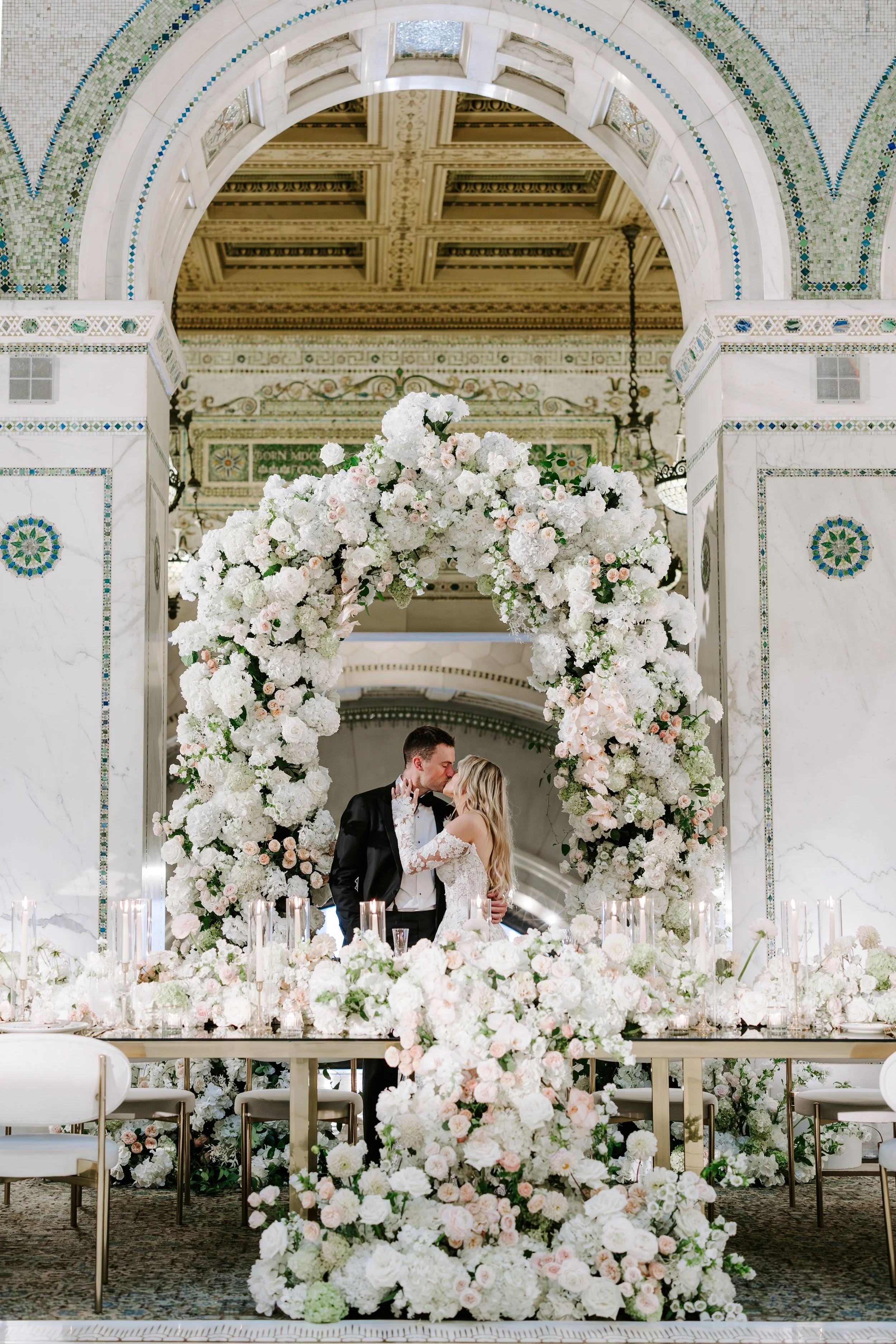 A bride and groom kissing under a floral arch at their wedding ceremony, with candles and floral arrangements on the table in front of them.