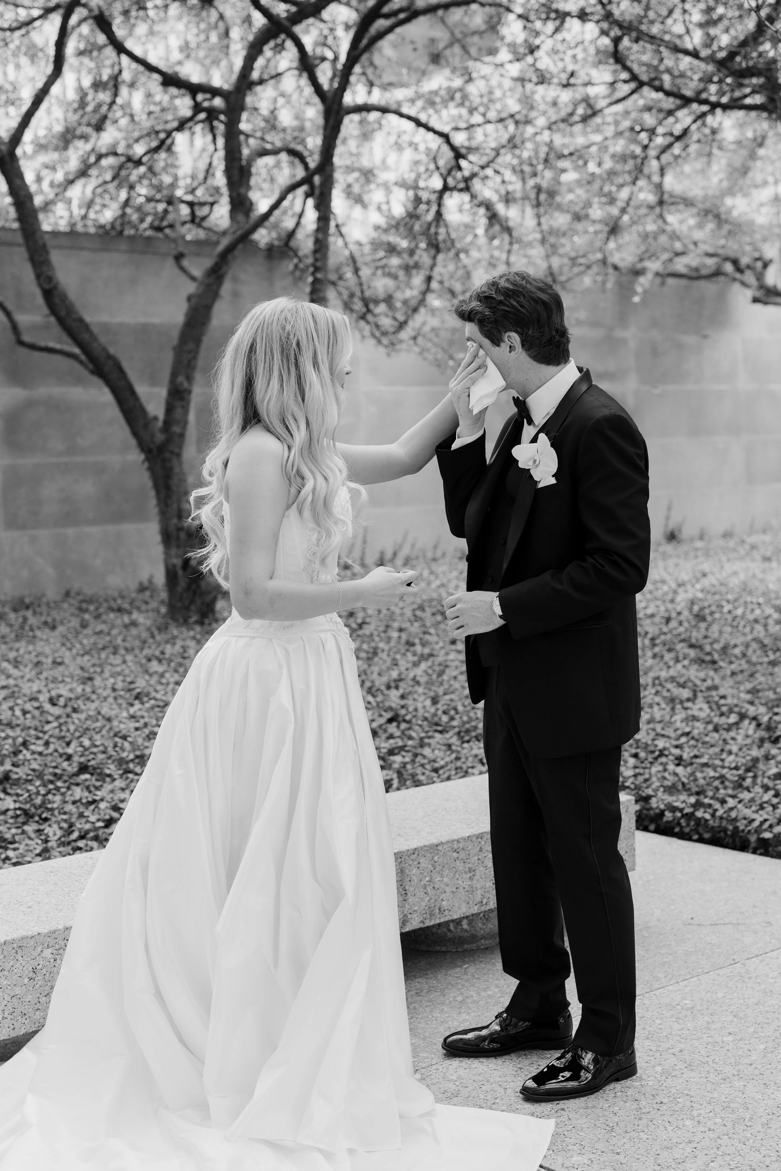 A bride with long blonde hair in a white wedding dress stands outside, touching a groom's face, who is dressed in a black tuxedo with a bow tie and a white boutonniere, as he wipes tears with a tissue; a tree and a wall are visible in the background.