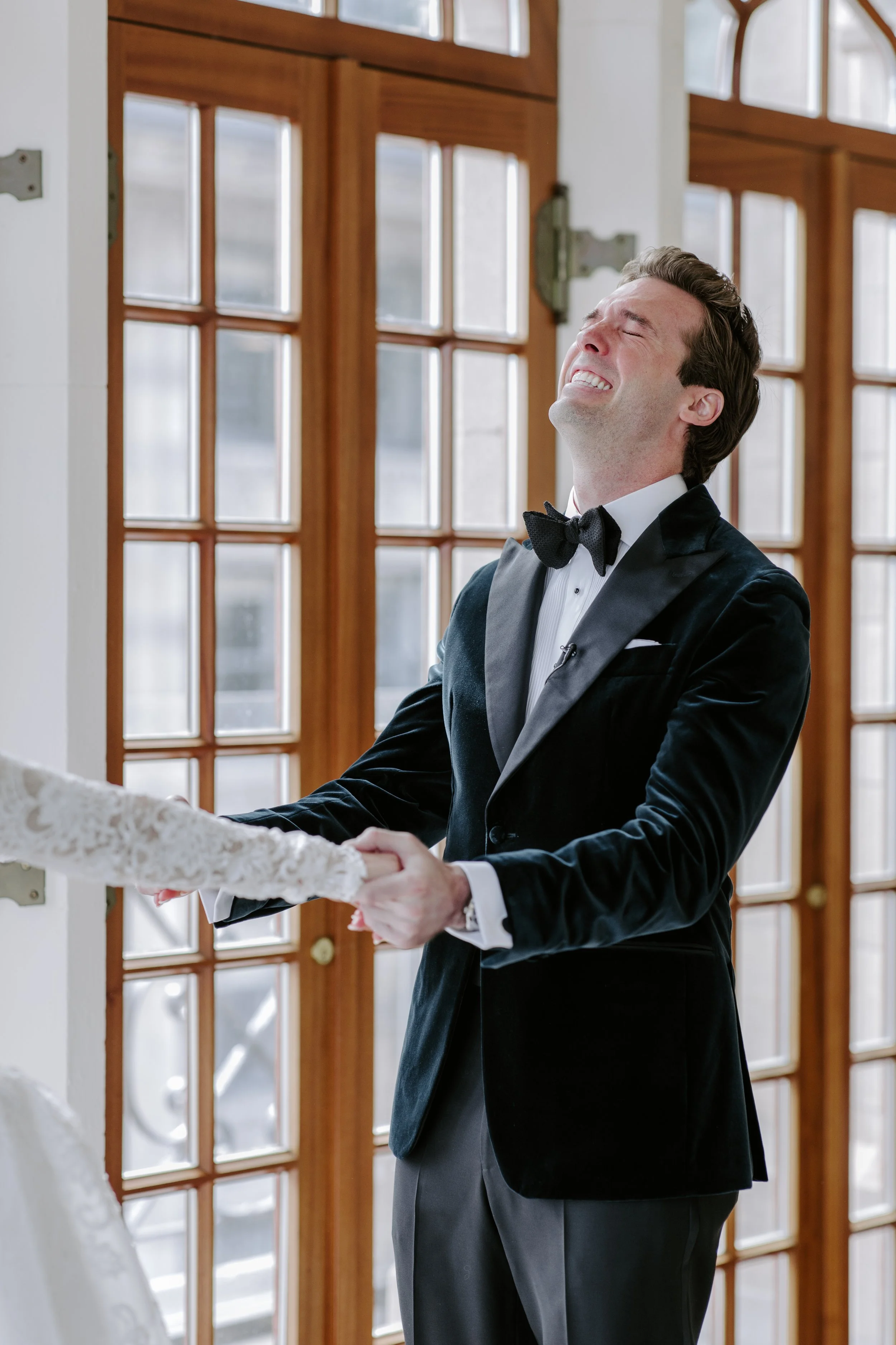 A man in a tuxedo is smiling and holding hands with a woman in a white lace dress during a wedding ceremony indoors in front of large glass doors.