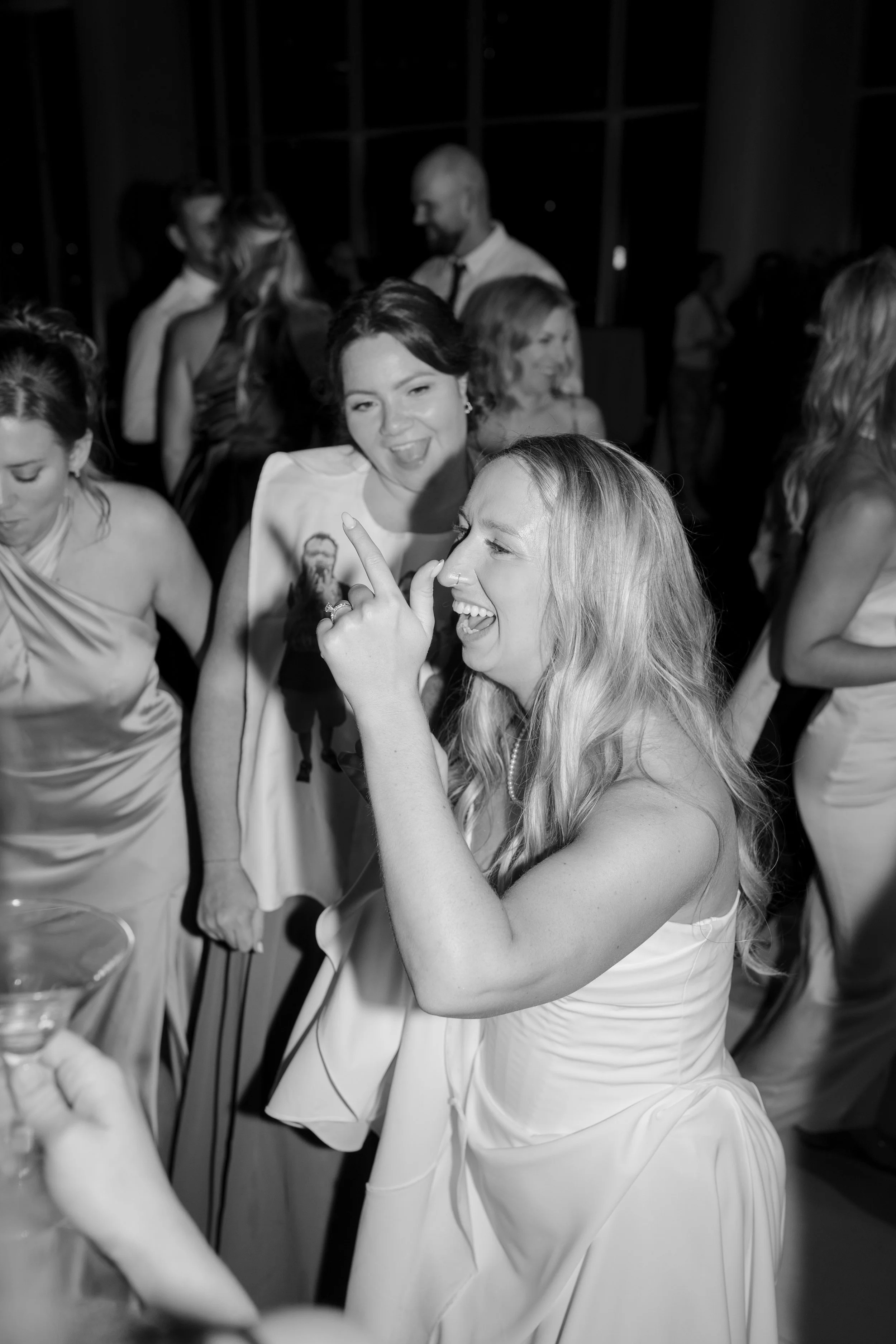Group of women in formal dresses at a celebration, with a woman in the foreground smiling and pointing, and other women and a man in background.