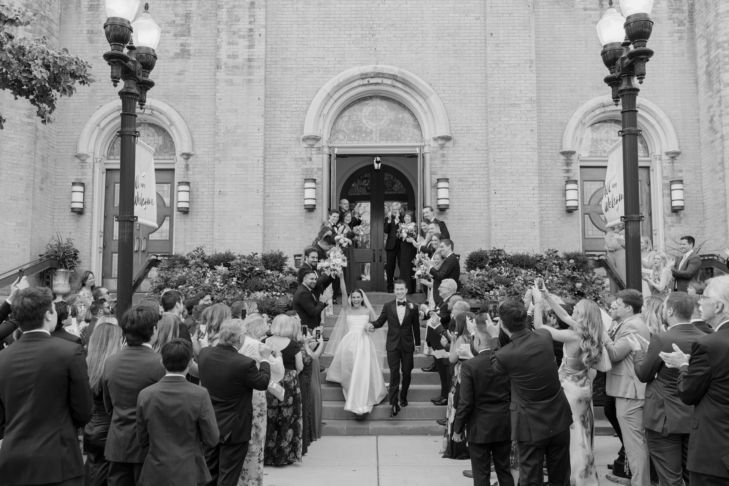 A wedding celebration with a bride and groom walking down stairs outside a church, surrounded by guests. The guests are clapping and taking photos, and the church has brick walls and large arched doors.