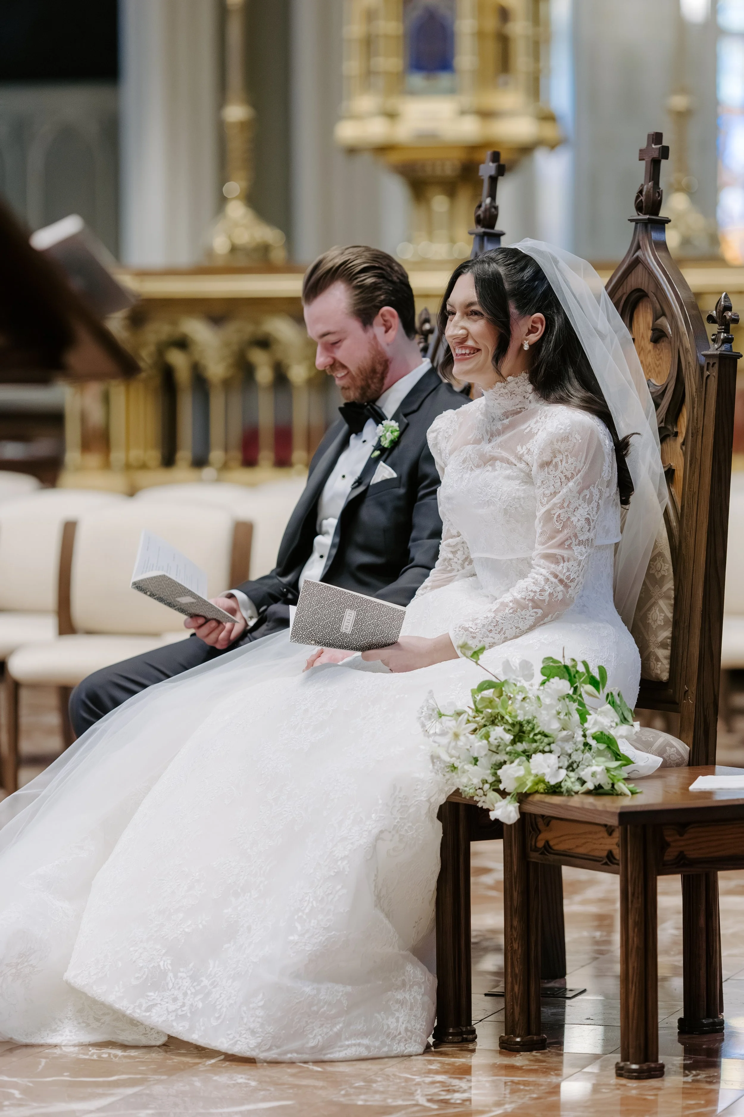 A bride and groom sitting on church pews during their wedding ceremony, smiling and reading prayer books. The bride is wearing a white lace wedding dress and veil, holding a bouquet of white flowers. The groom is in a black tuxedo, with a white shirt