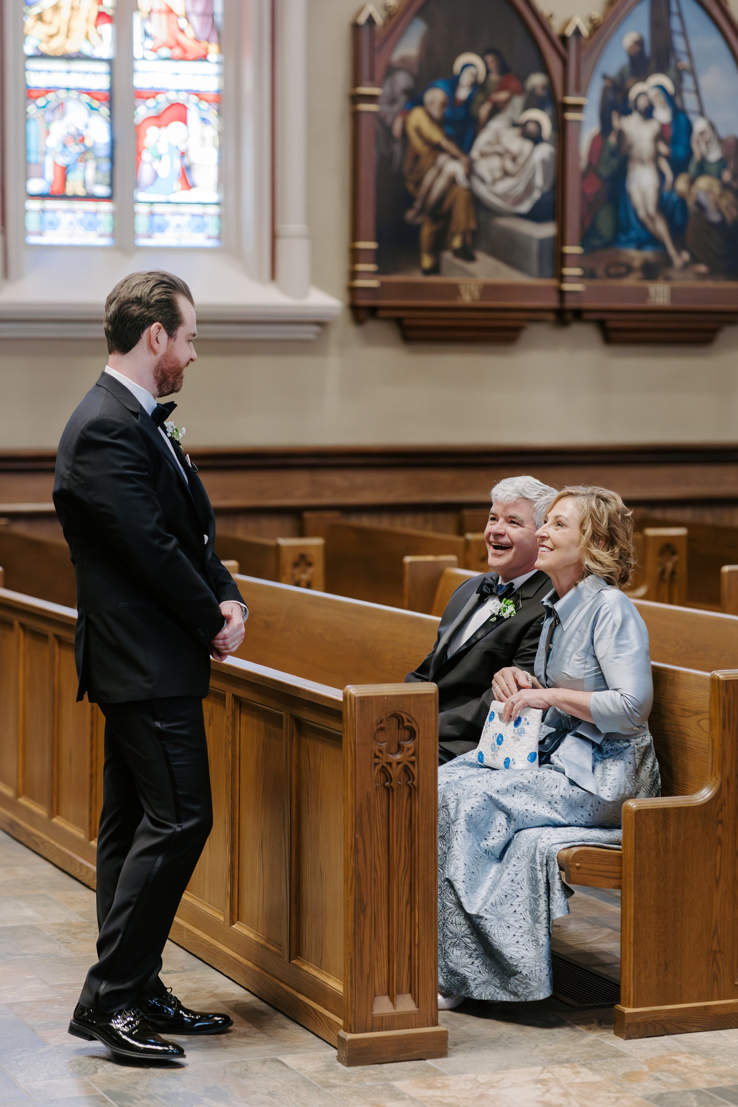 A groom in a black tuxedo and shiny black shoes is standing in front of a church pew, looking at a couple sitting inside the pew. The couple is smiling and looking at the groom. The church has stained glass windows and religious paintings on the wall