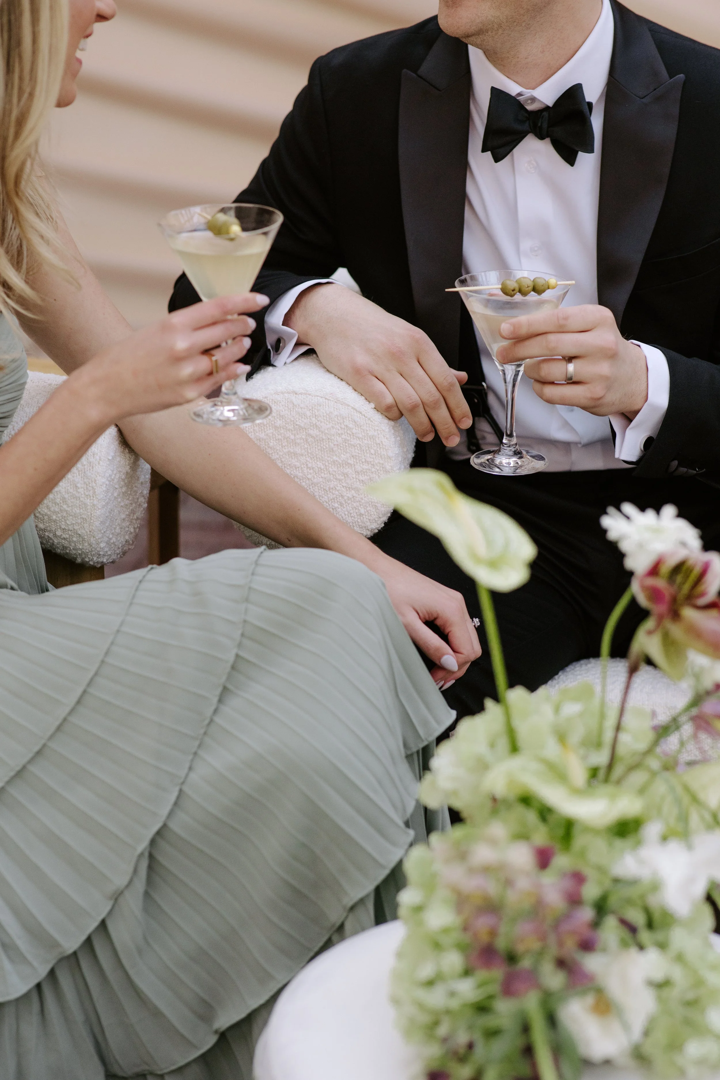 A woman and a man holding martini glasses with green olives, dressed in formal attire, celebrating at an elegant event with floral arrangements in the foreground.
