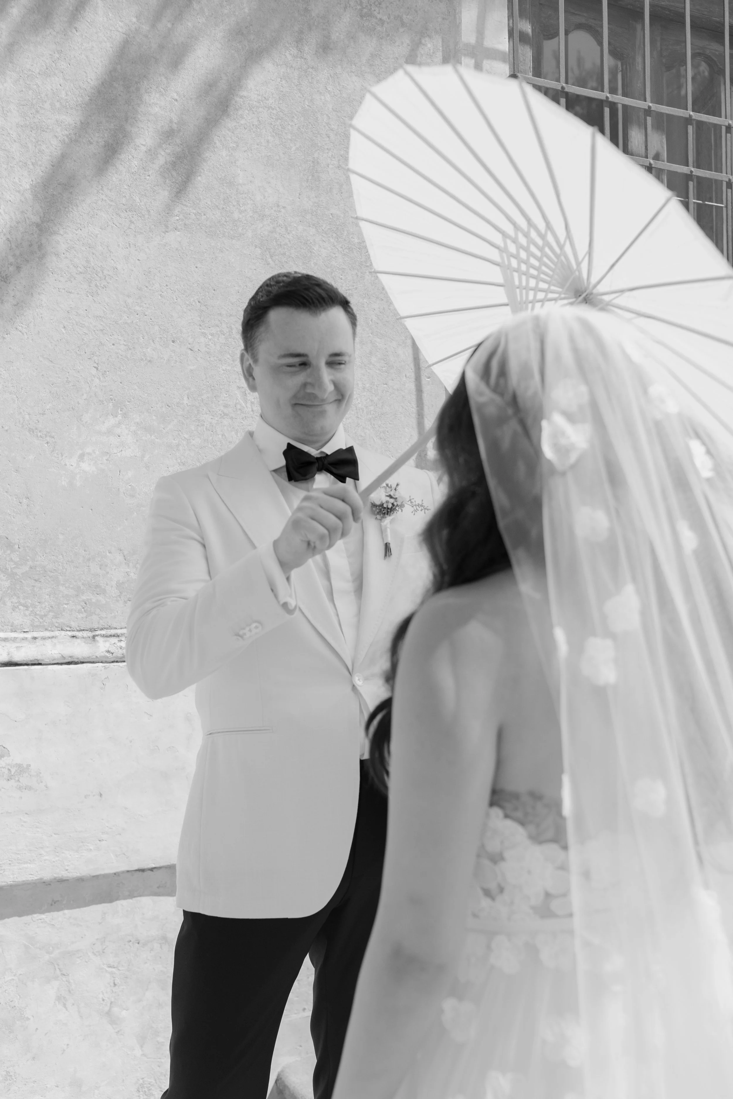 A groom in a white tuxedo with a black bow tie holding a white umbrella, smiling at a bride with dark hair and a veil, standing outside near a textured wall and a building with a balcony.