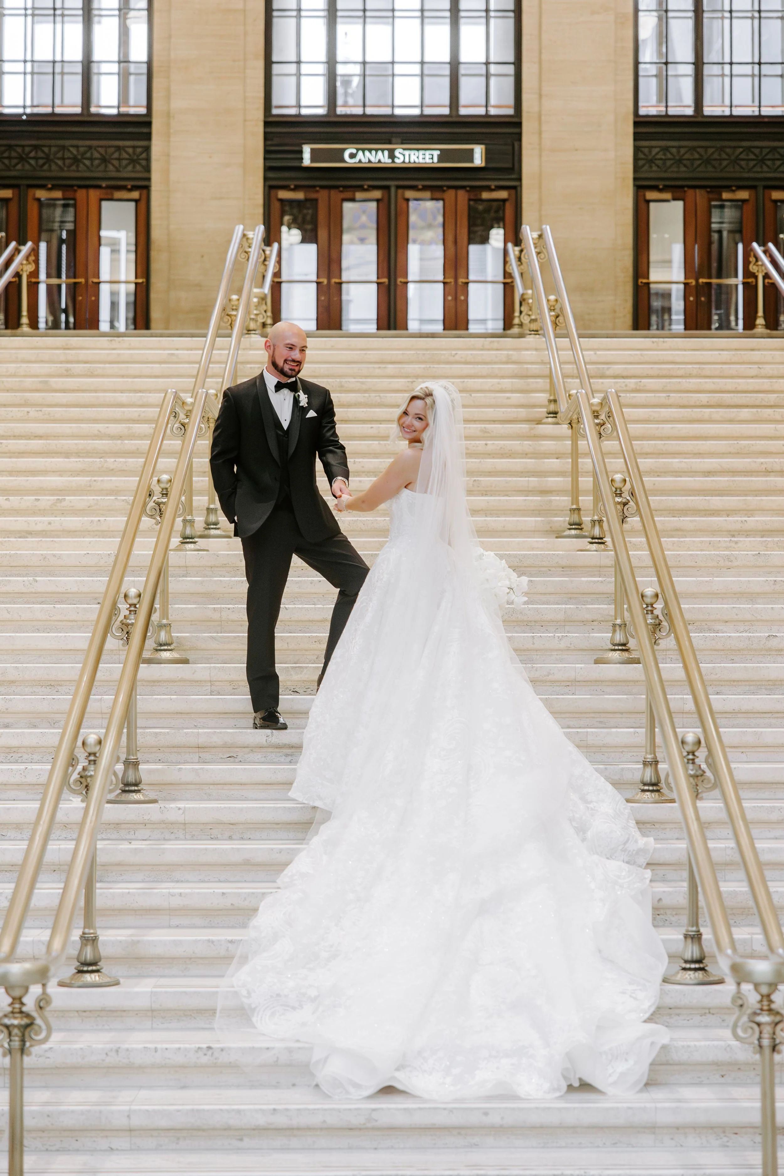 A bride and groom holding hands and smiling on a grand staircase inside a building labeled 'Canal Street'.