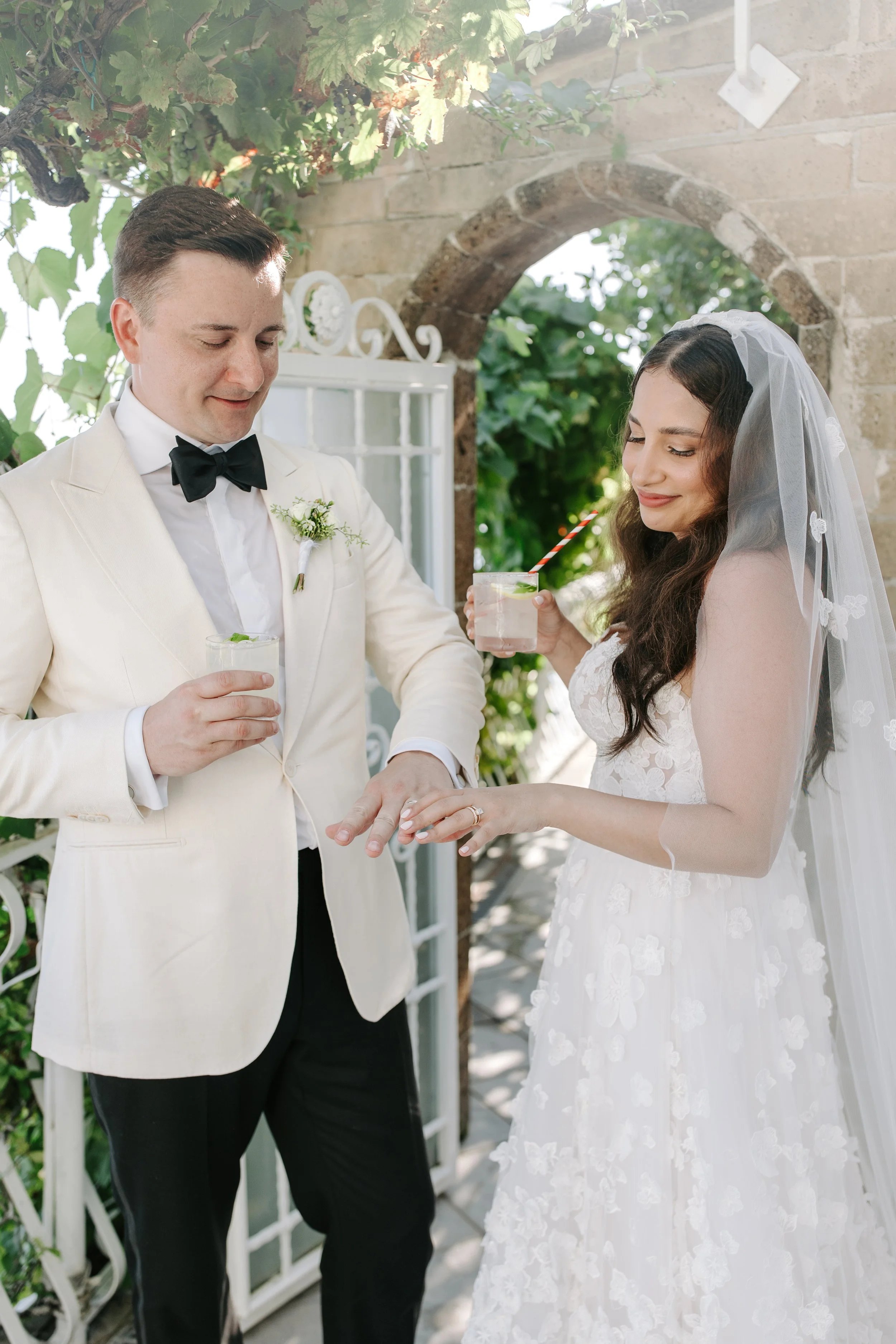 A bride and groom at their wedding ceremony exchanging rings outdoors, with the groom wearing a cream tuxedo and the bride in a white lace gown with a veil, holding a drink with a striped straw.