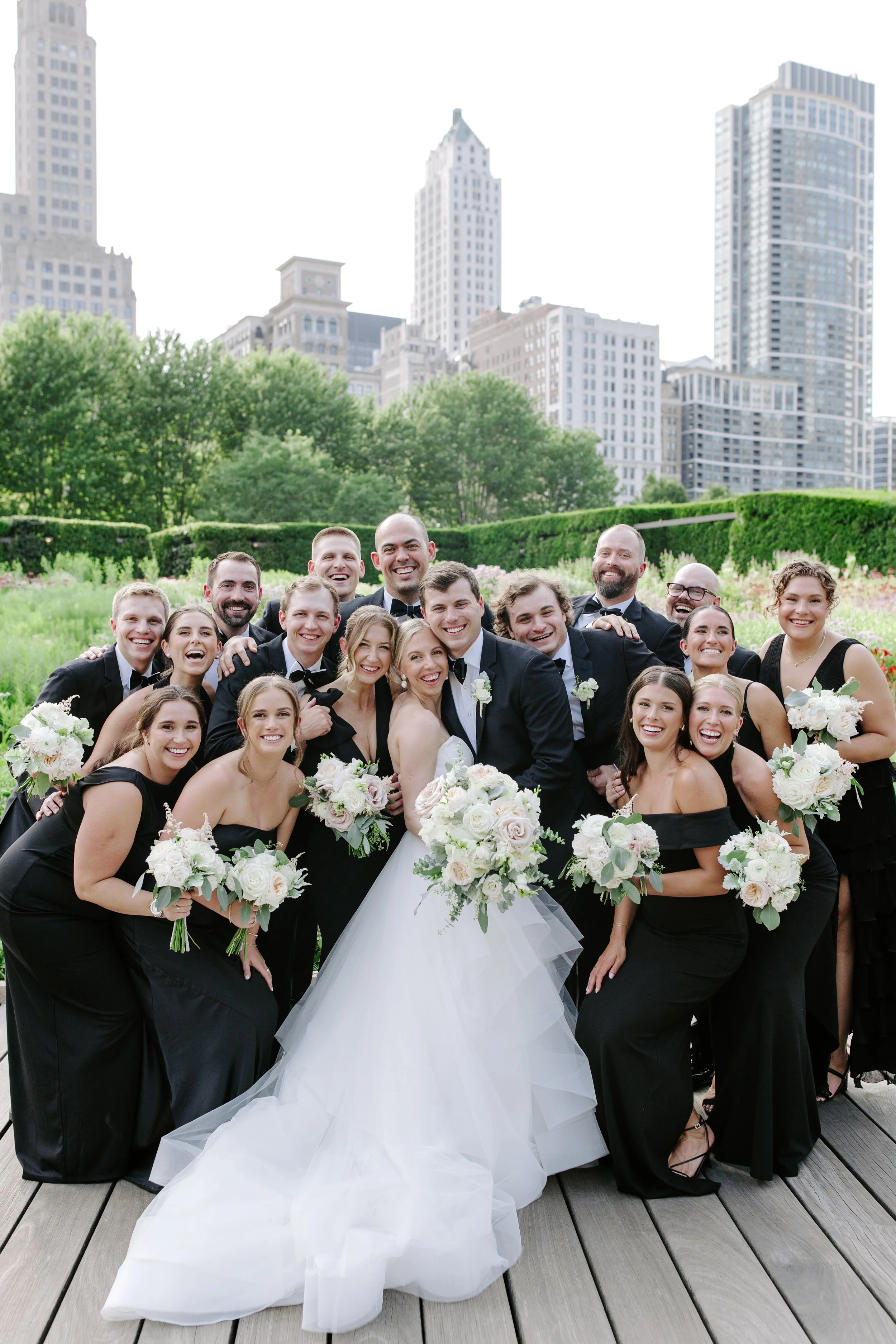 A large group of wedding guests, including men in tuxedos and women in black dresses, gather outdoors on a wooden deck in front of greenery and cityscape buildings. The bride in a white wedding gown is holding a bouquet, smiling and surrounded by fri